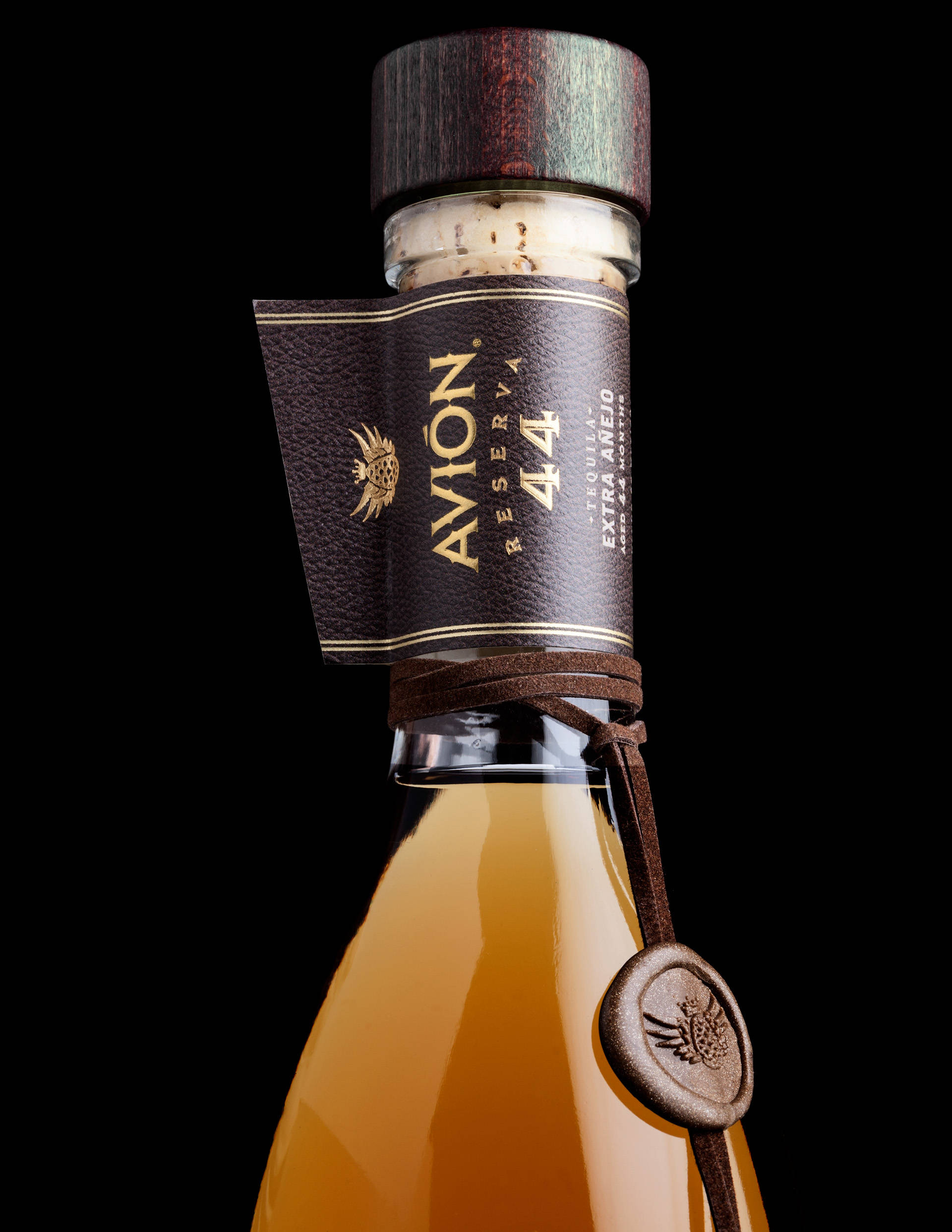 bottle 桌布 壁紙 — Avion Reserva 44 龍舌蘭酒瓶特寫，琥珀色液體、皮革標籤和木蓋，黑色背景。