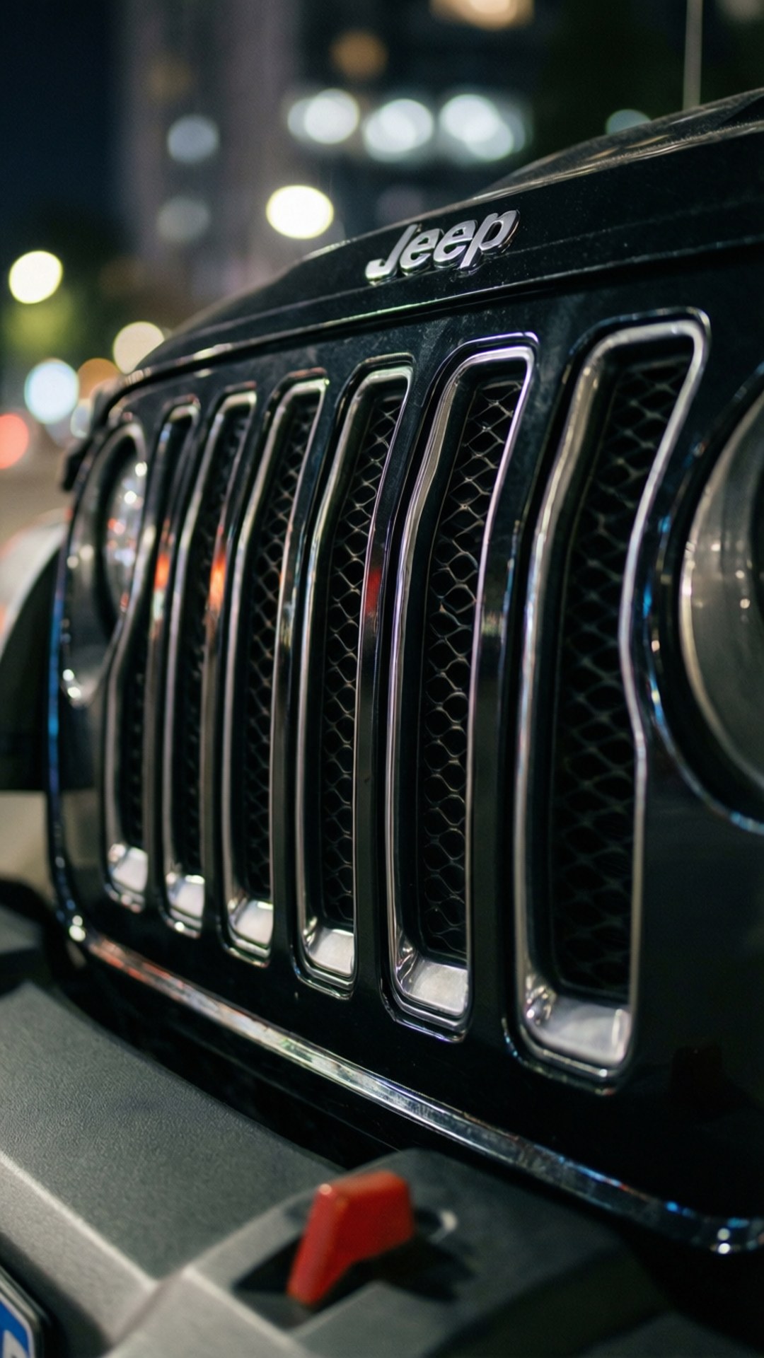 Grille Detail Night 壁紙 — 黑色Jeep水箱護罩特寫，帶有鍍鉻飾邊和紅色拖車鉤，背景是夜間模糊的城市燈光。