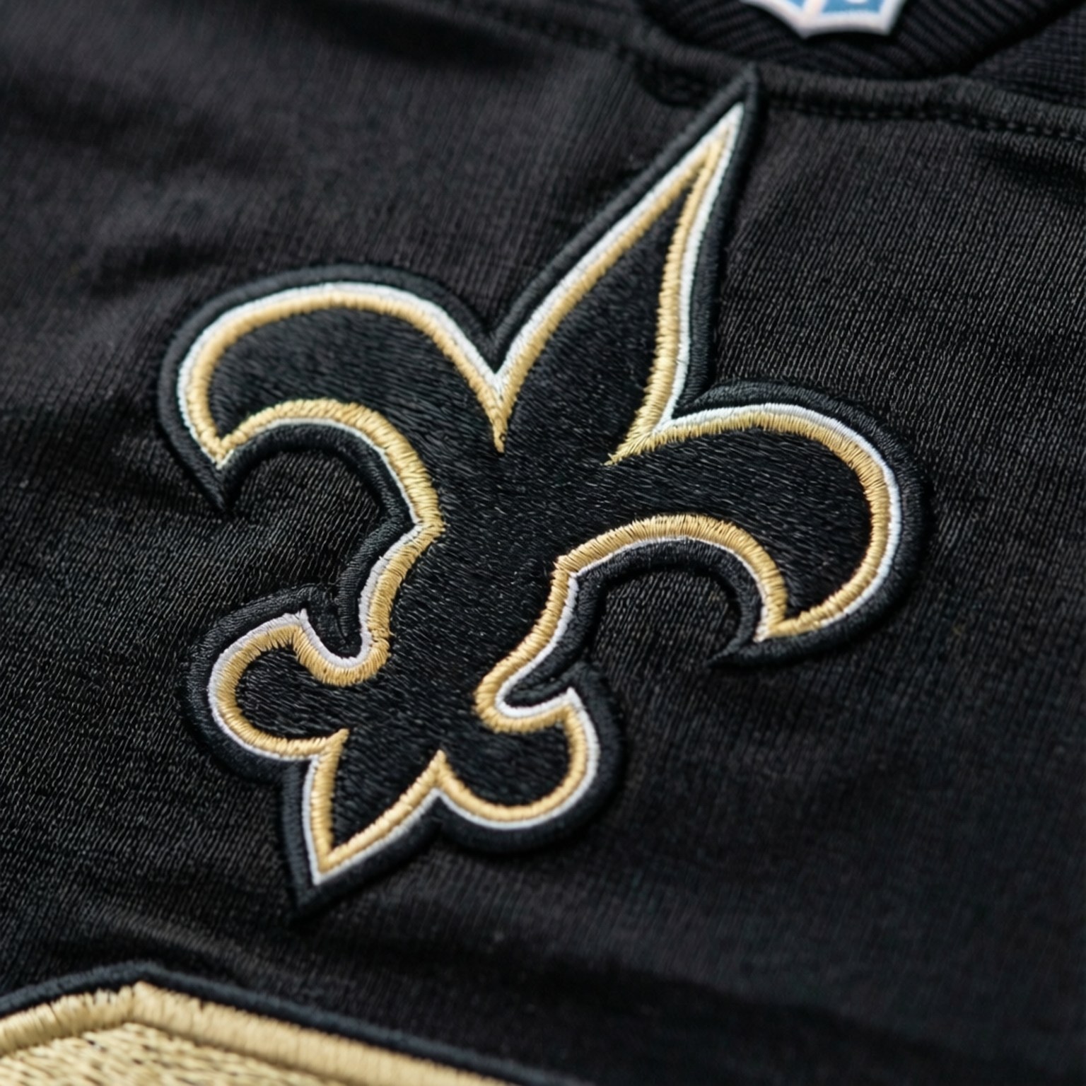 Saints Jersey Detail 壁紙 — 黑色運動球衣的特寫，帶有金色和白色線精細刺繡的鳶尾花標誌。