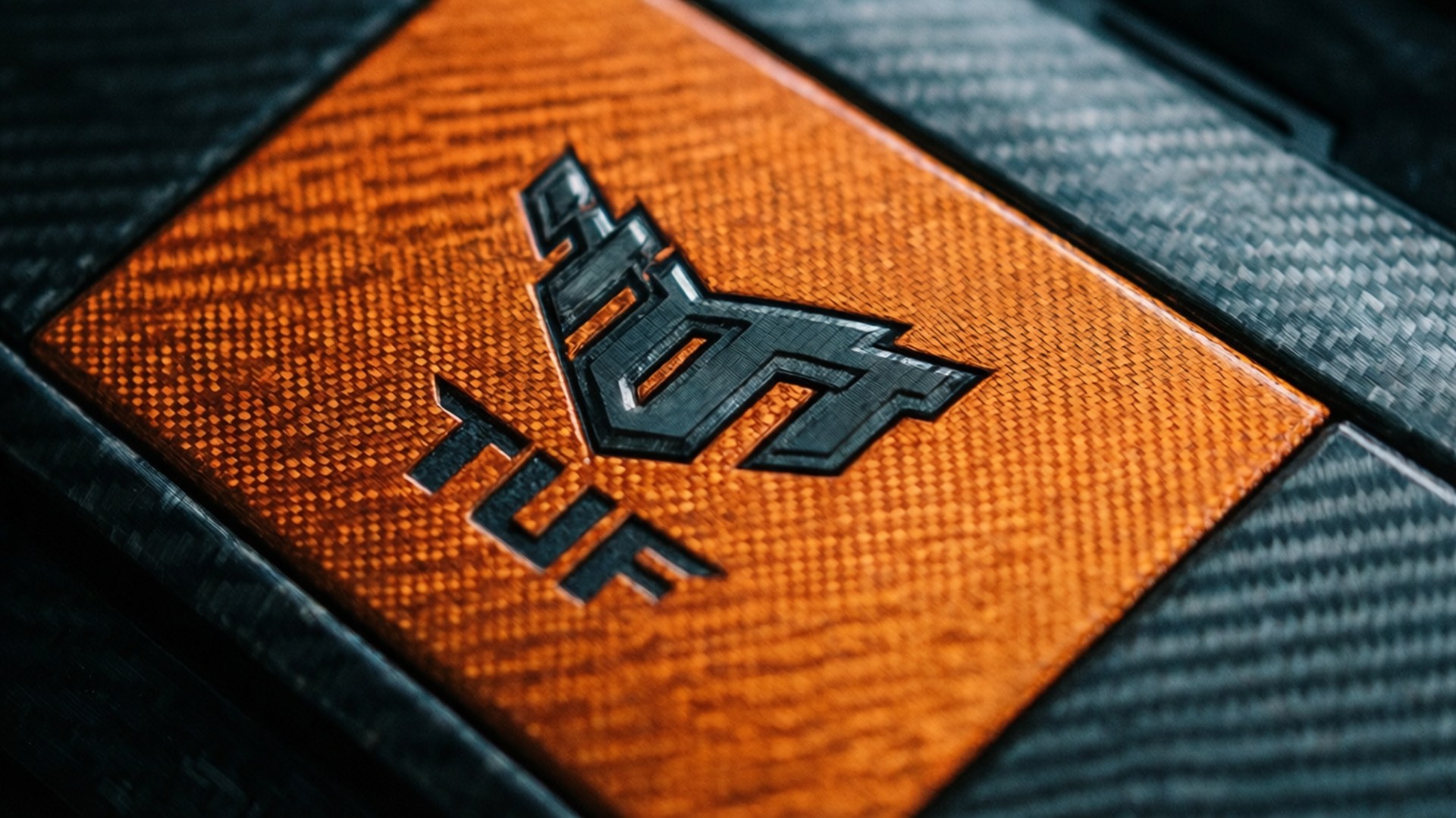 Carbon Fiber TUF 壁紙 — 近距離拍攝一個大膽的黑色TUF標誌，壓印在帶有紋理的橙色和碳纖維表面上。