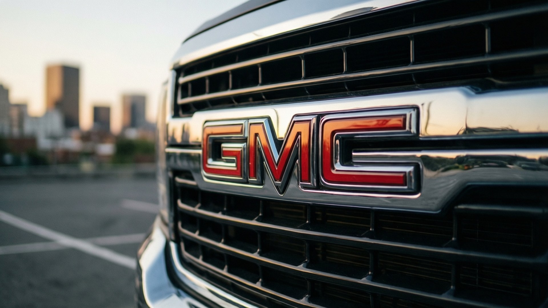 GMC Grille Close-up 壁紙 — 近距離拍攝卡車格柵上的 GMC 標誌，背景是日落時分模糊的城市天際線。