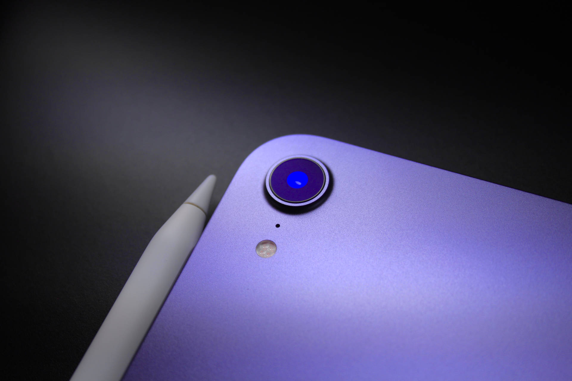 紫色iPad，相機鏡頭發出藍光，旁邊是白色Apple Pencil。 — iPad 壁紙