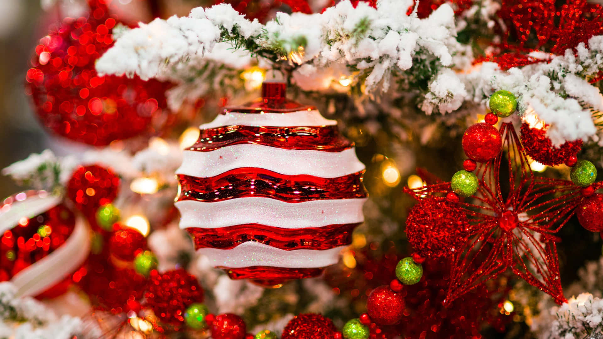  壁紙 — 一個紅白條紋玻璃裝飾品的特寫，掛在灑滿雪的聖誕樹上，樹上點綴著閃亮的紅色、綠色和金色裝飾。