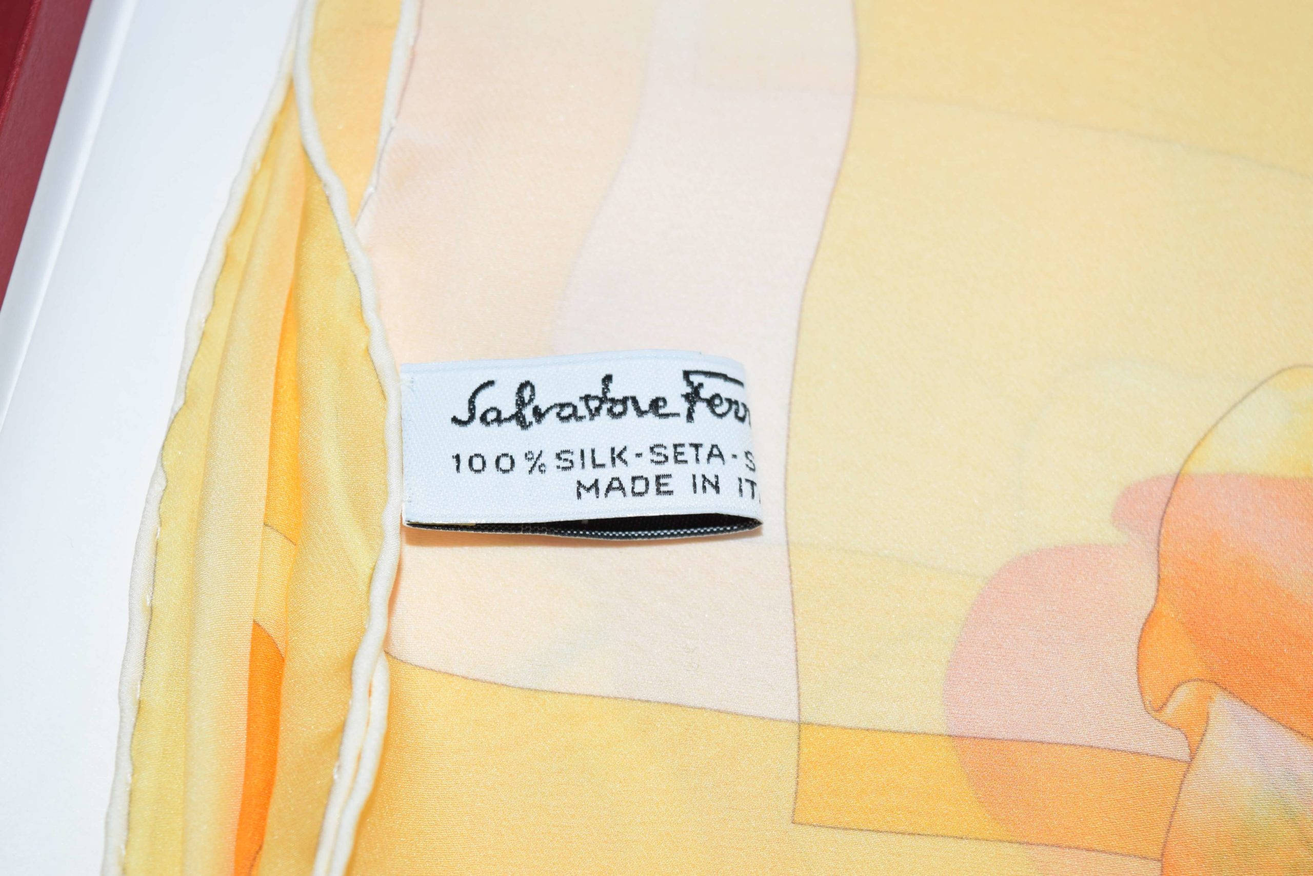 Ferragamo 桌布 壁紙 — Salvatore Ferragamo 絲巾標籤的特寫，顯示100%絲綢材質和義大利製造，背景為柔和的黃色抽象圖案。