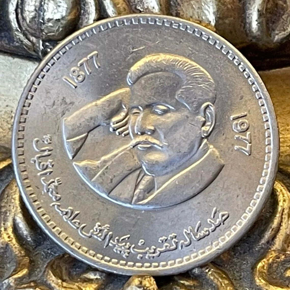  壁紙 — 銀幣特寫，上面有男性肖像、阿拉伯文字，以及刻有1877年和1977年，放置在帶紋理的金色金屬上。