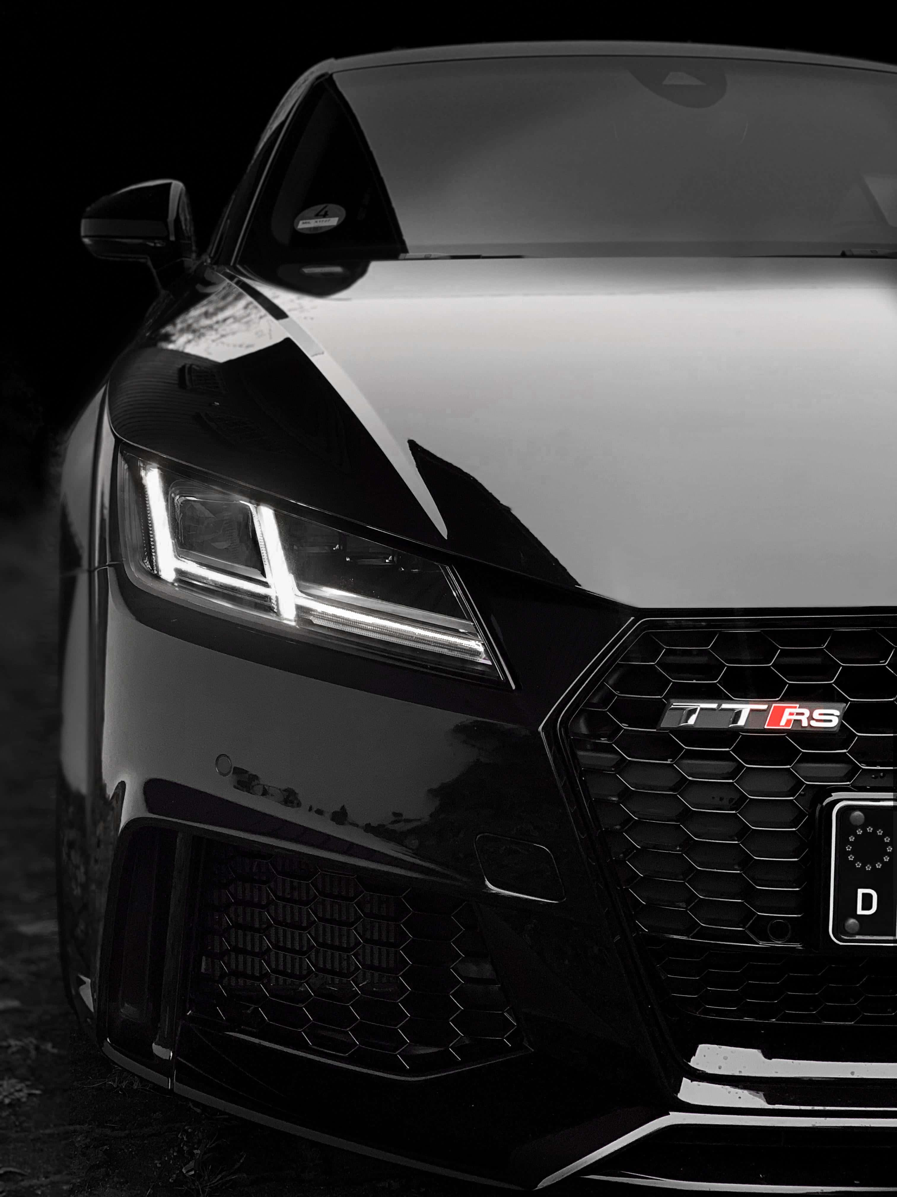 Audi Tt Rs 桌布 壁紙 — 黑色奧迪 TT RS 跑車特寫，配備發光頭燈和六角形水箱護罩，背景漆黑。