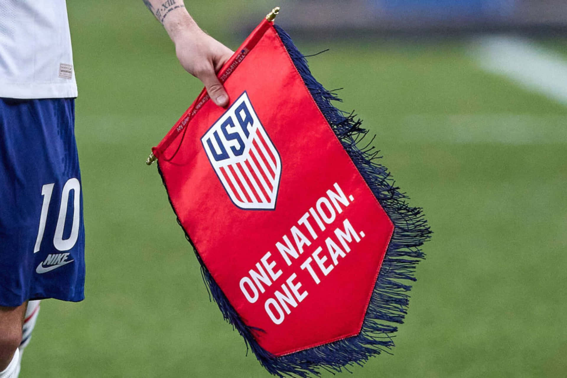  壁紙 — 一名足球員手持帶有流蘇的紅色美國國旗，上面有口號 one nation one team 的特寫。