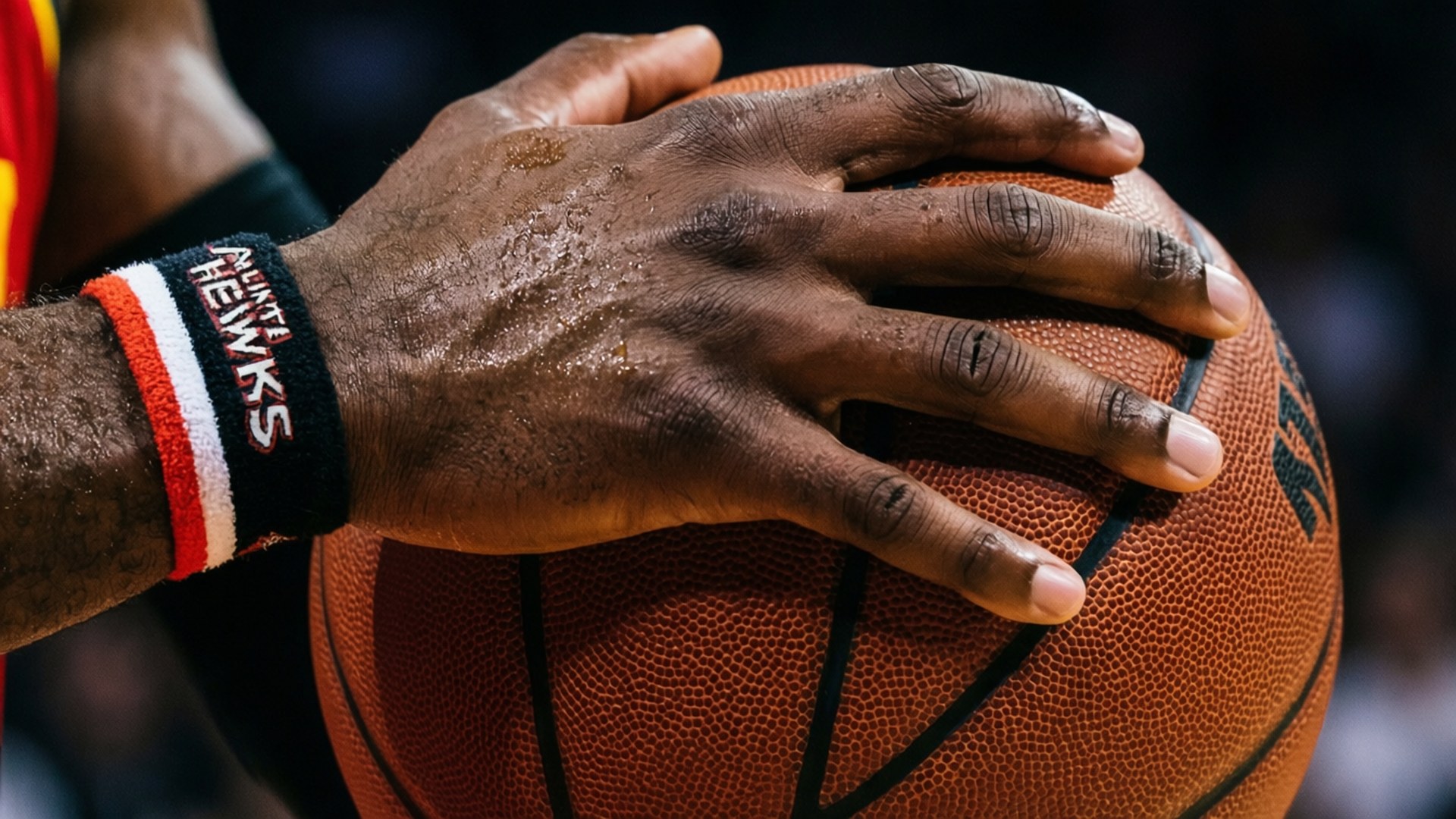 Grip and Wristband 壁紙 — 一個近距離拍攝，汗流浹背的籃球運動員的手緊握著一個帶有黑色線條和「HAWKS」腕帶的橙色球。