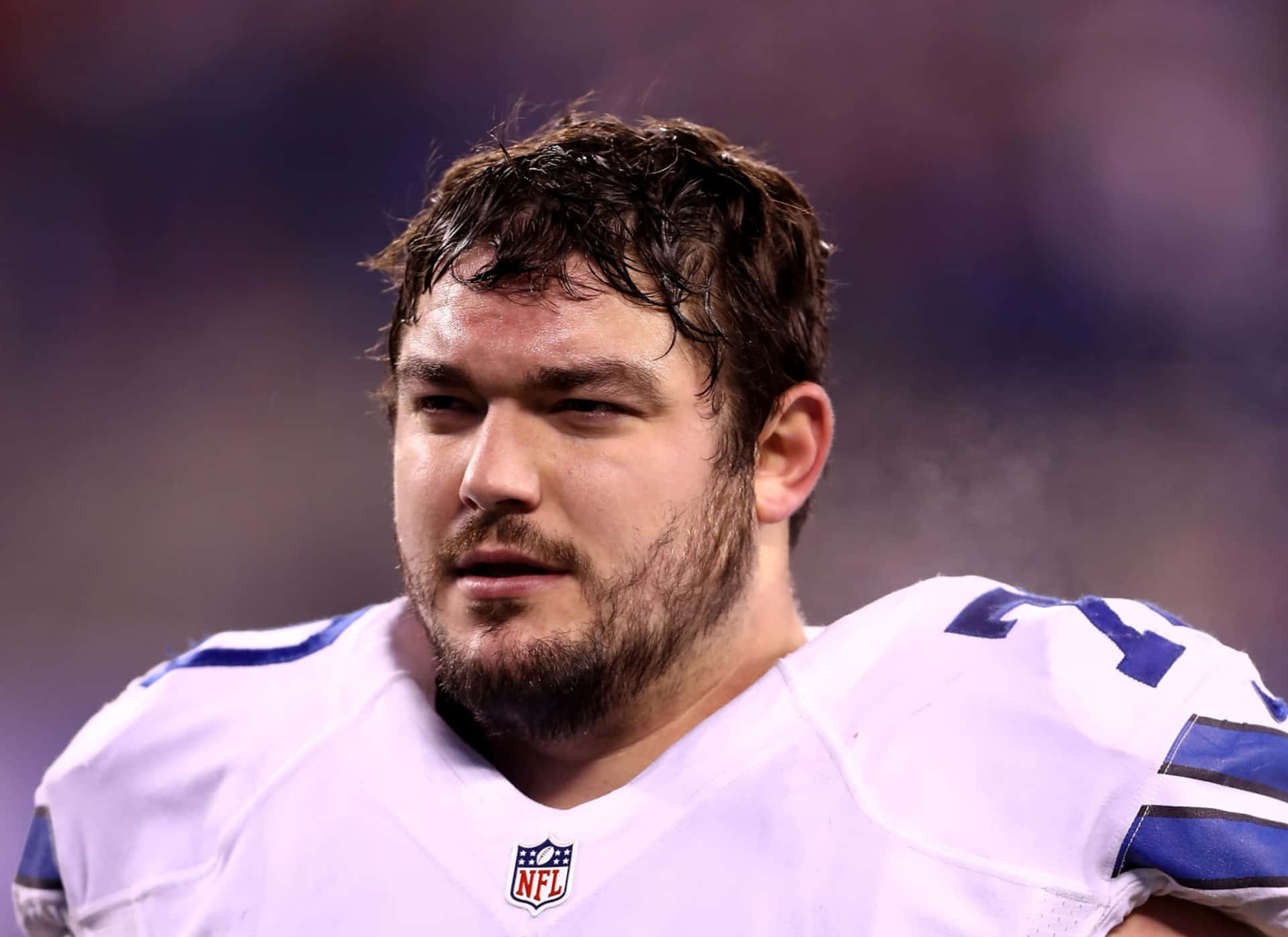  壁紙 — NFL 美式足球員身穿白色和藍色球衣，在球場燈光下比賽時汗流浹背的近距離特寫，表情激烈。