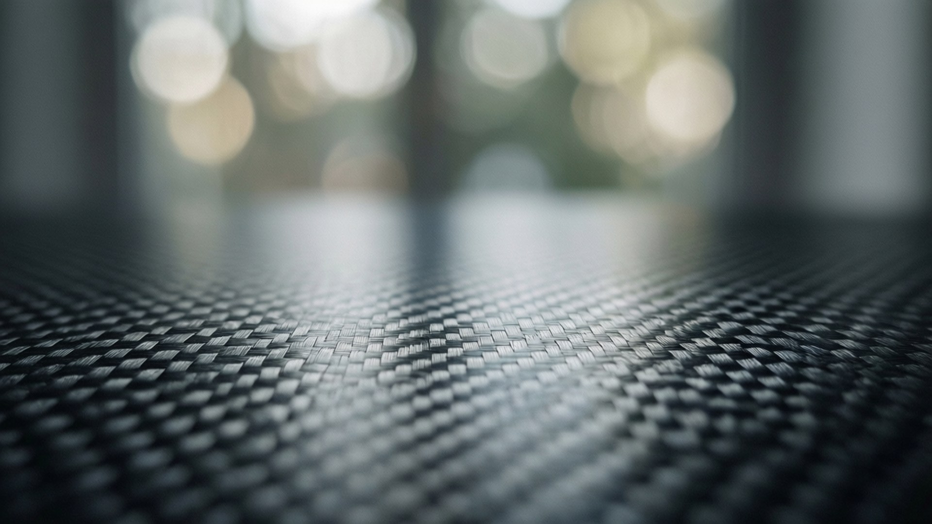 Soft Focus Carbon Fiber Background 壁紙 — 黑色紋理碳纖維表面的特寫，背景有柔和模糊的金色散景燈光。