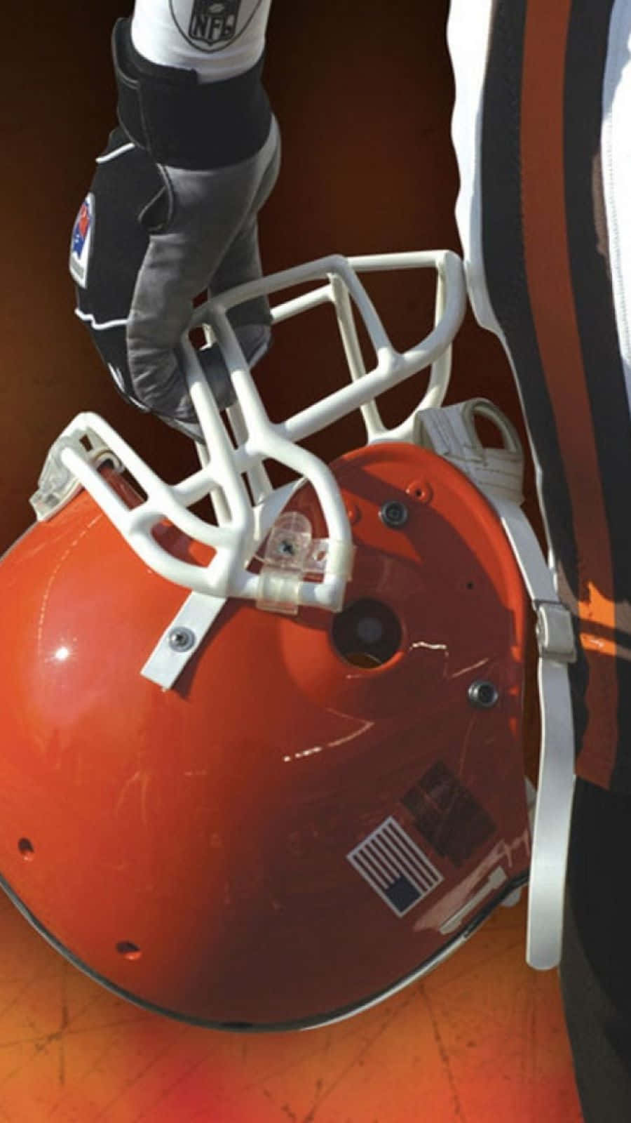  壁紙 — 一名身穿球衣的球員手持克里夫蘭布朗隊NFL美式足球頭盔的特寫，背景為溫暖的橙色。