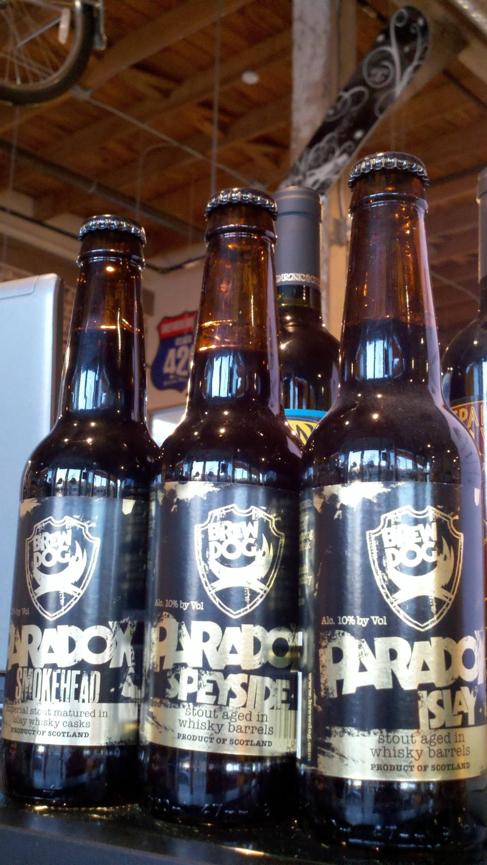 Drink 桌布 壁紙 — BrewDog Paradox 黑啤酒玻璃瓶的特寫，排列在木樑下的架子上，上方有一個自行車輪。