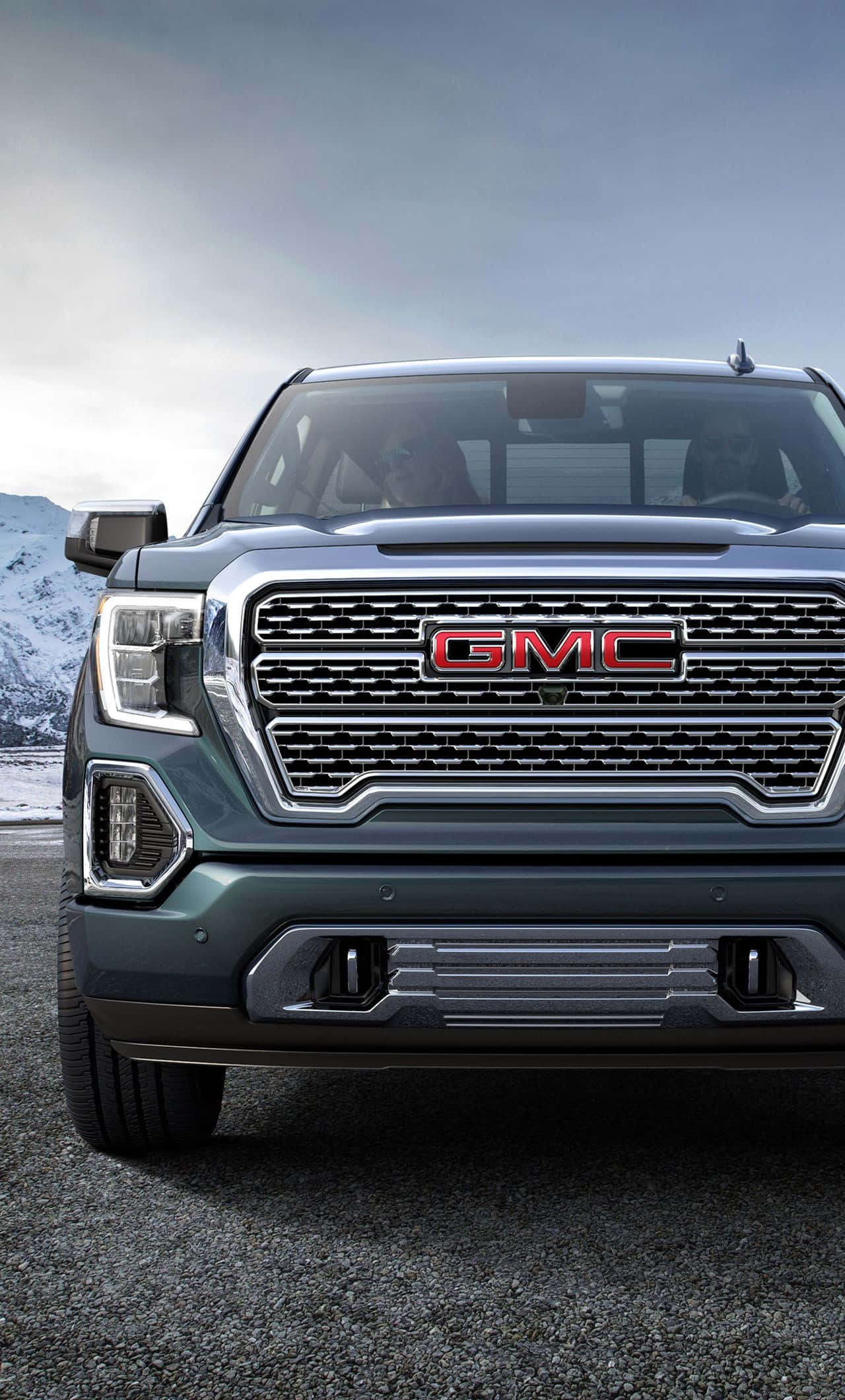 Gmc Yukon 桌布 壁紙 — 特寫鏡頭：深綠色GMC卡車的前面格柵，帶有紅色標誌，背景是陰天下的雪山。