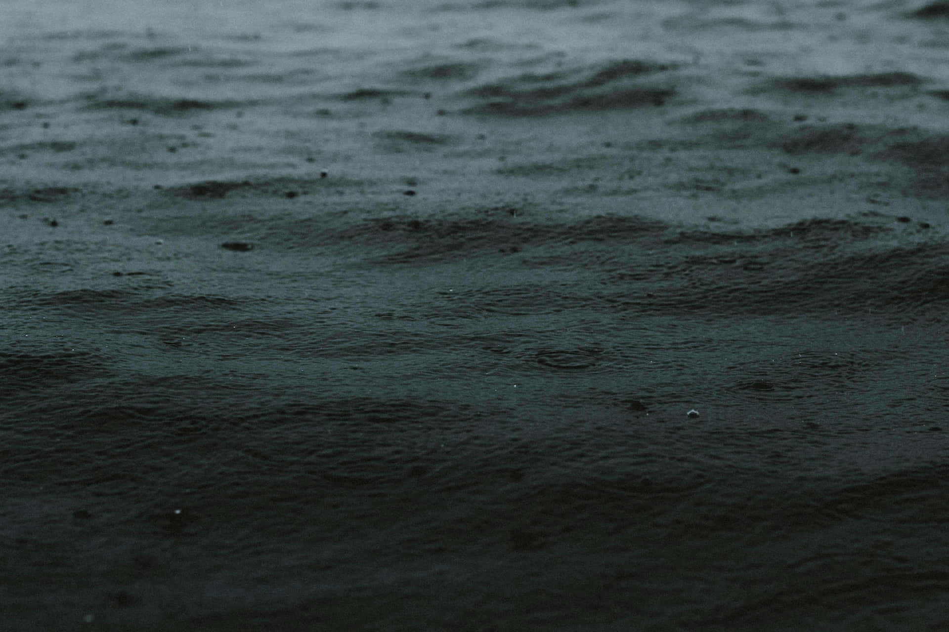  壁紙 — 昏暗光線下深色漣漪水面的特寫，雨滴在水面形成細小漣漪。
