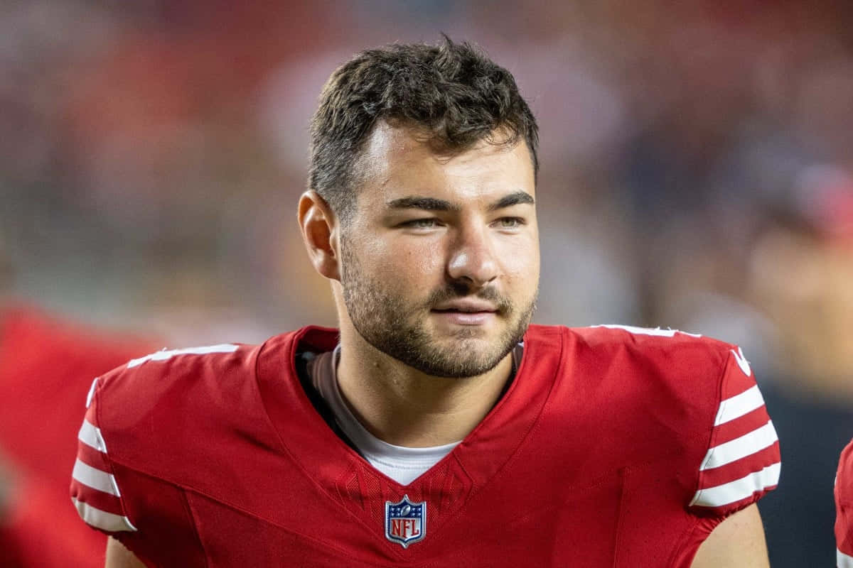  壁紙 — 專注的NFL美式足球員身穿紅色帶白色條紋球衣，在比賽中望向鏡頭外的特寫。