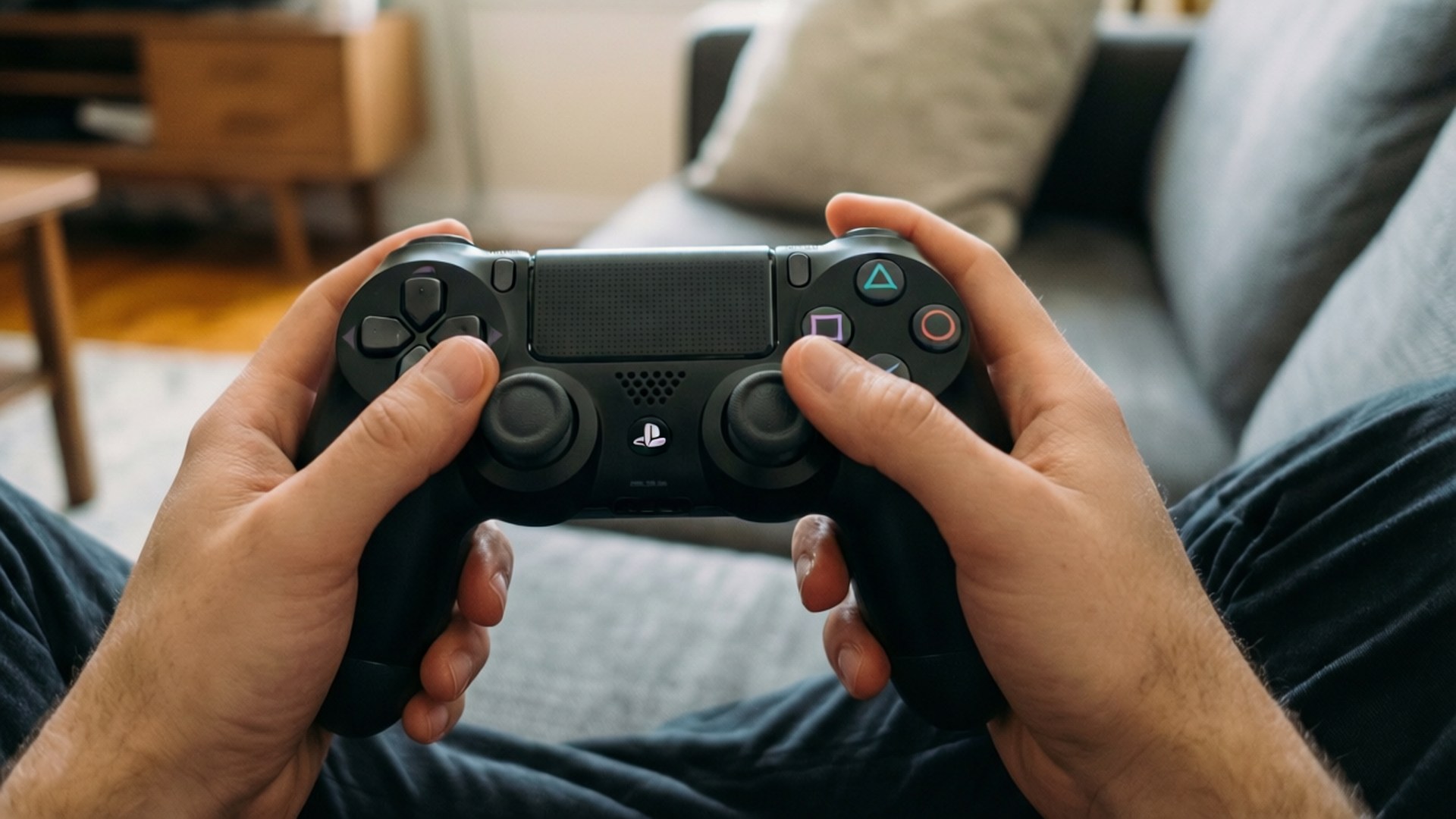 Gamer's View PS4 壁紙 — 特寫雙手緊握黑色 PlayStation 手把，坐在舒適客廳的灰色沙發上。