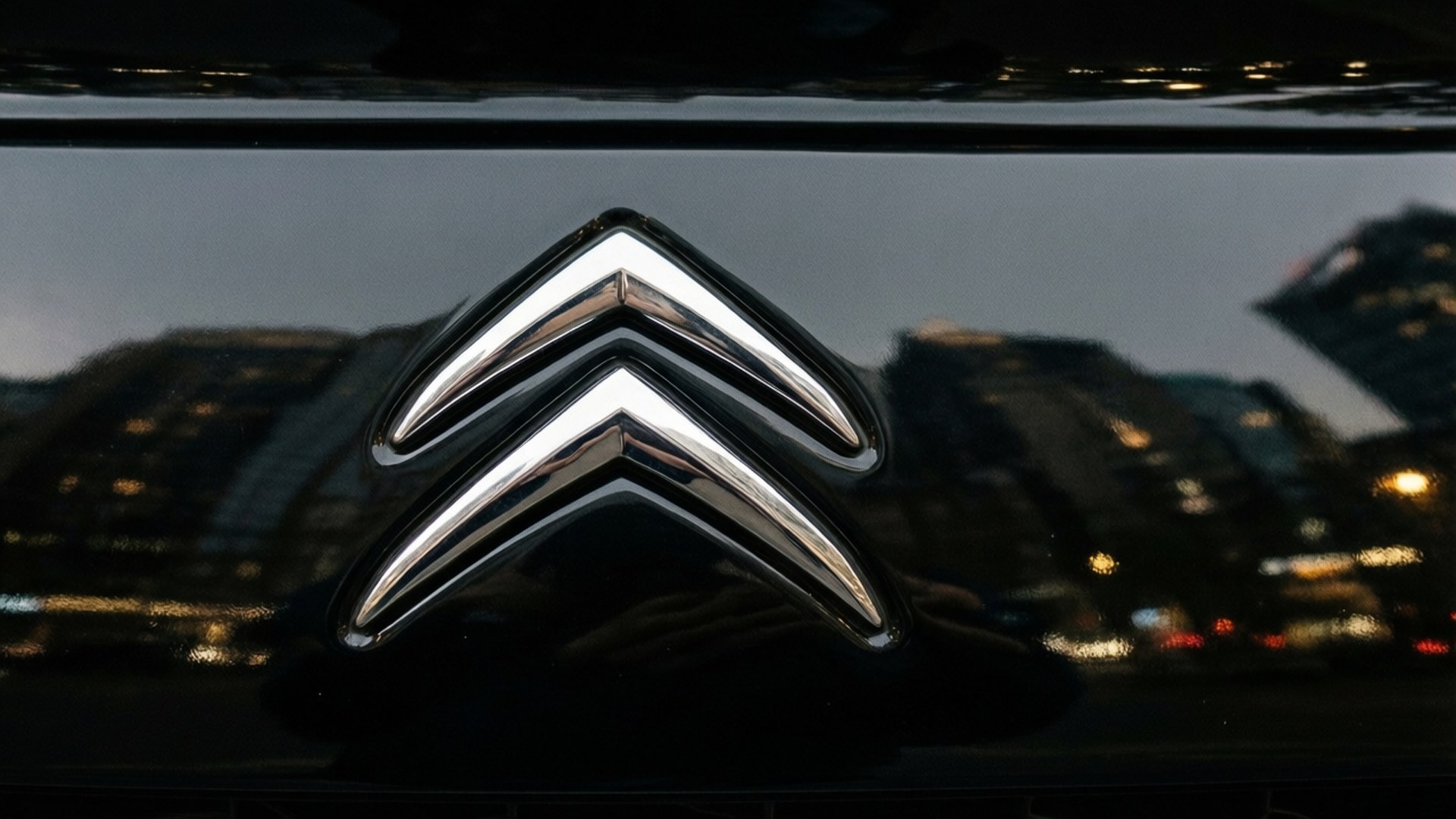 Citroen Logo Macro 壁紙 — 黑色引擎蓋上閃亮鍍鉻雪鐵龍標誌的特寫，映照出黃昏時模糊的城市景觀。