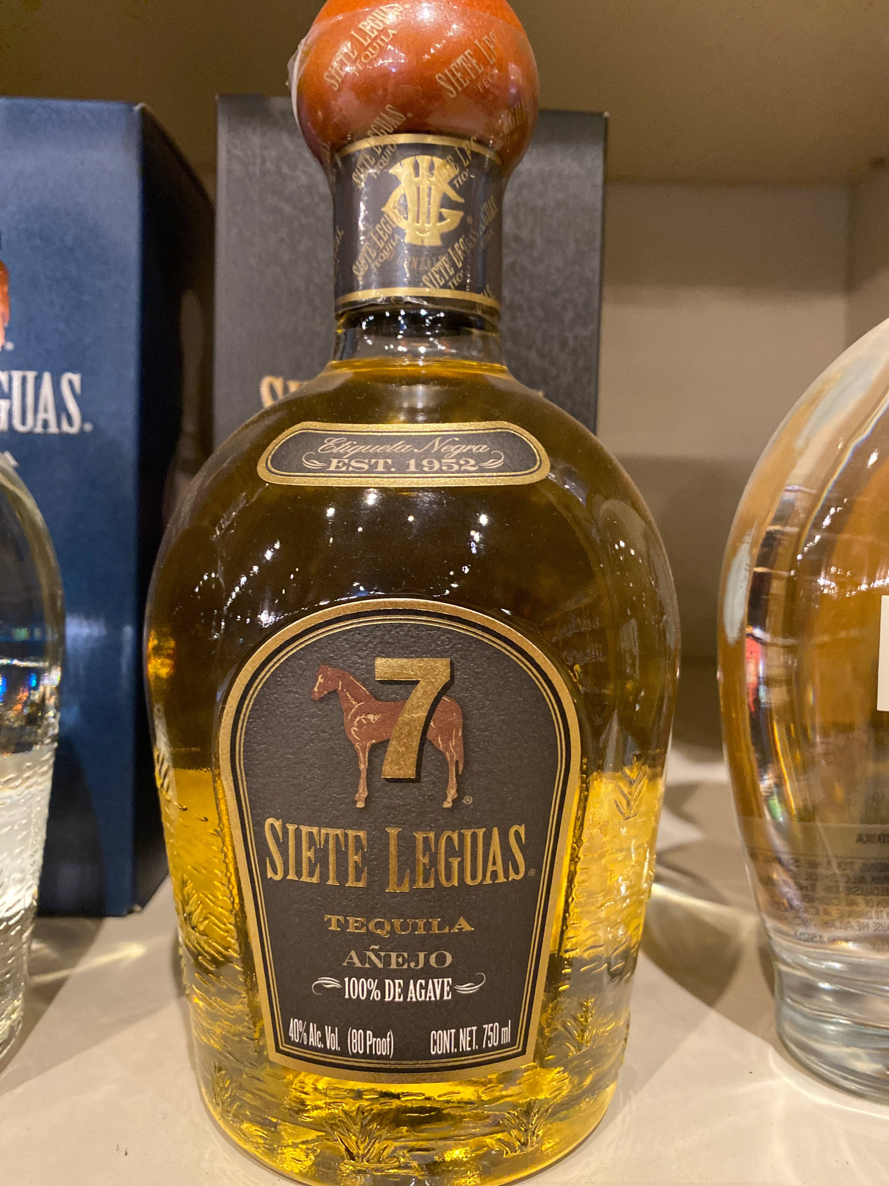 iPhone 桌布 壁紙 — Siete Leguas Añejo 龍舌蘭酒瓶的特寫，瓶內有琥珀色液體和華麗標籤，與其他烈酒一起陳列在架子上。