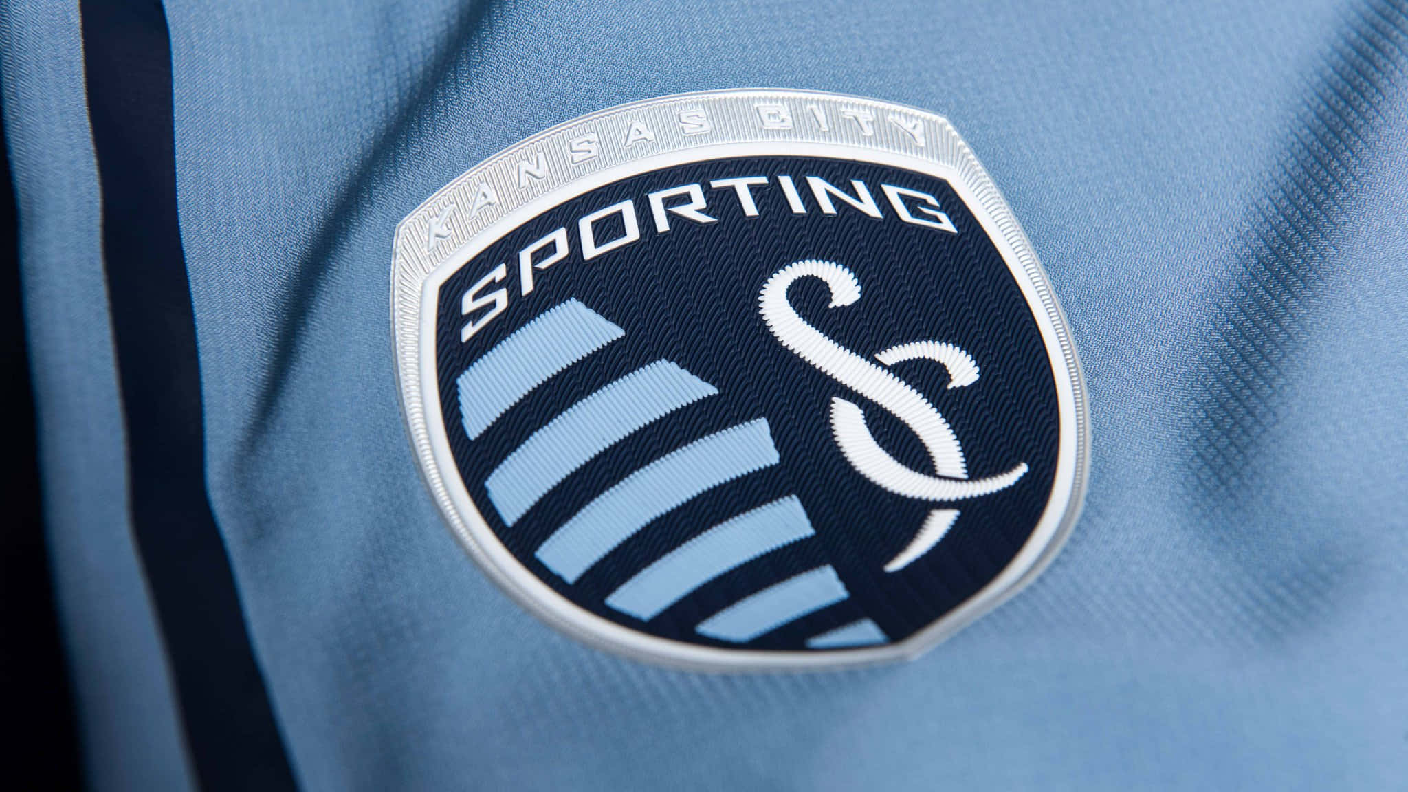 足球 桌布 壁紙 — Sporting Kansas City 足球隊徽章近距離特寫，繡在淺藍色運動球衣布料上。