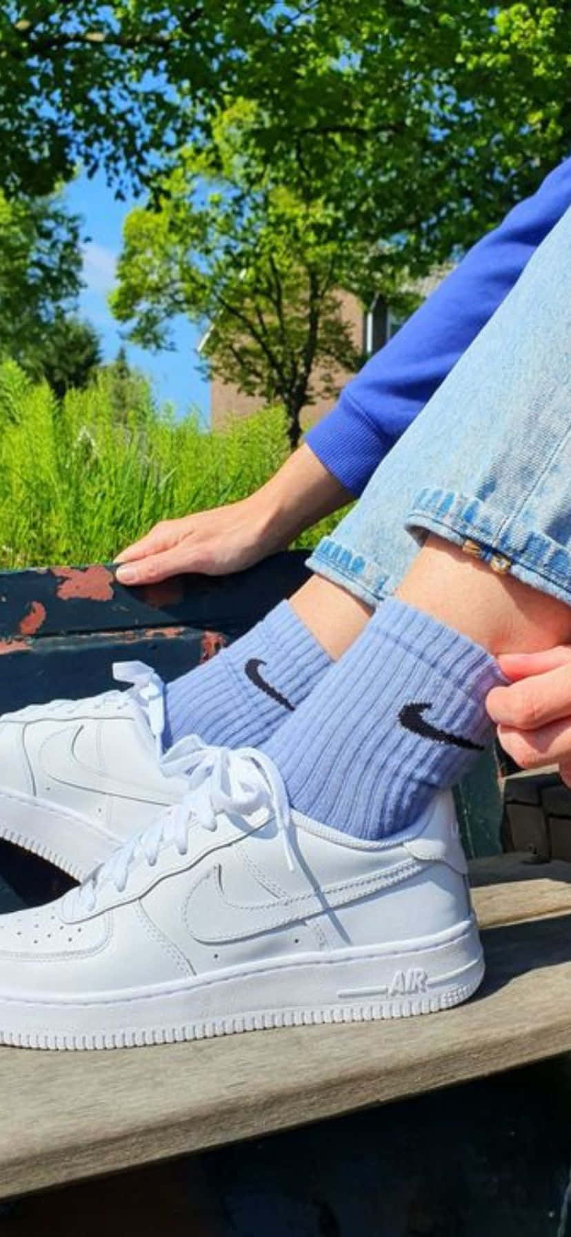  壁紙 — 特寫白色Nike Air Force 1運動鞋和淺藍色襪子放在木長椅上，上方是明亮的綠樹。