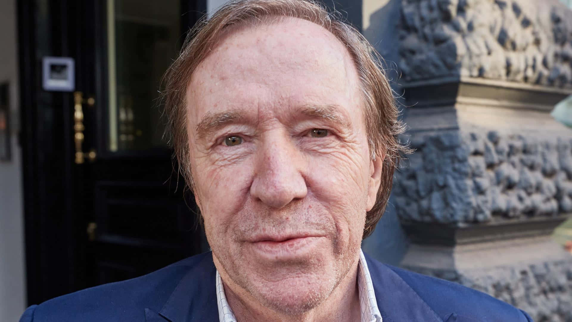  壁紙 — 中年男子的特寫肖像，他有棕色頭髮、穿著藍色西裝外套，站在室外的石紀念碑和黑色門附近。