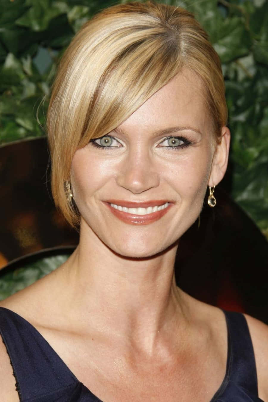 Natasha Henstridge 桌布 壁紙 — 一位微笑的金髮碧眼女子，身穿深藍色連衣裙，戴著金色耳環，以綠色樹葉為背景的特寫肖像。