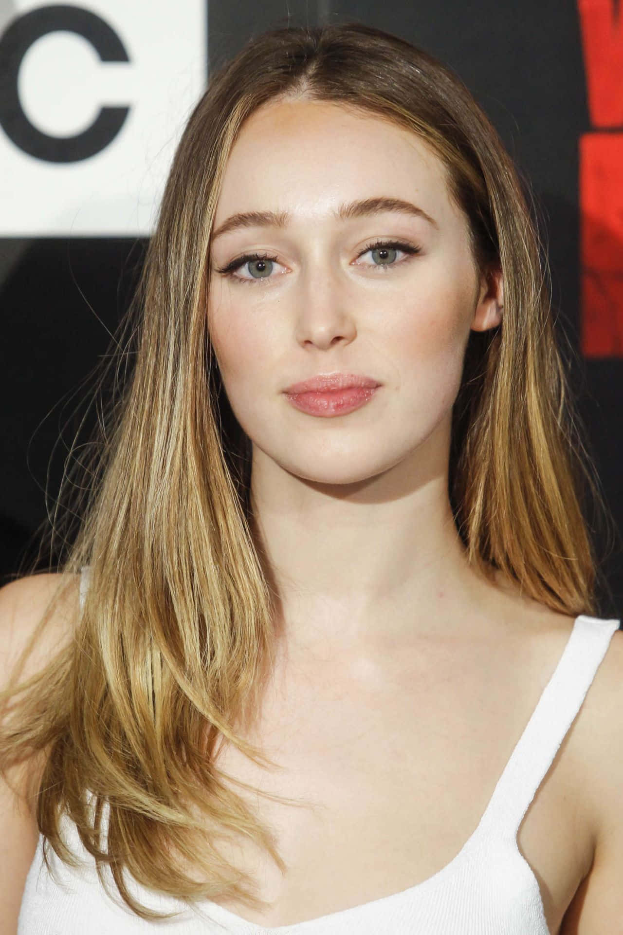 alycia debnam carey 桌布 壁紙 — 一位年輕女性的特寫肖像，長長的金髮和柔和的妝容，穿著白色背心，背景為帶有部分標誌的深色背景。