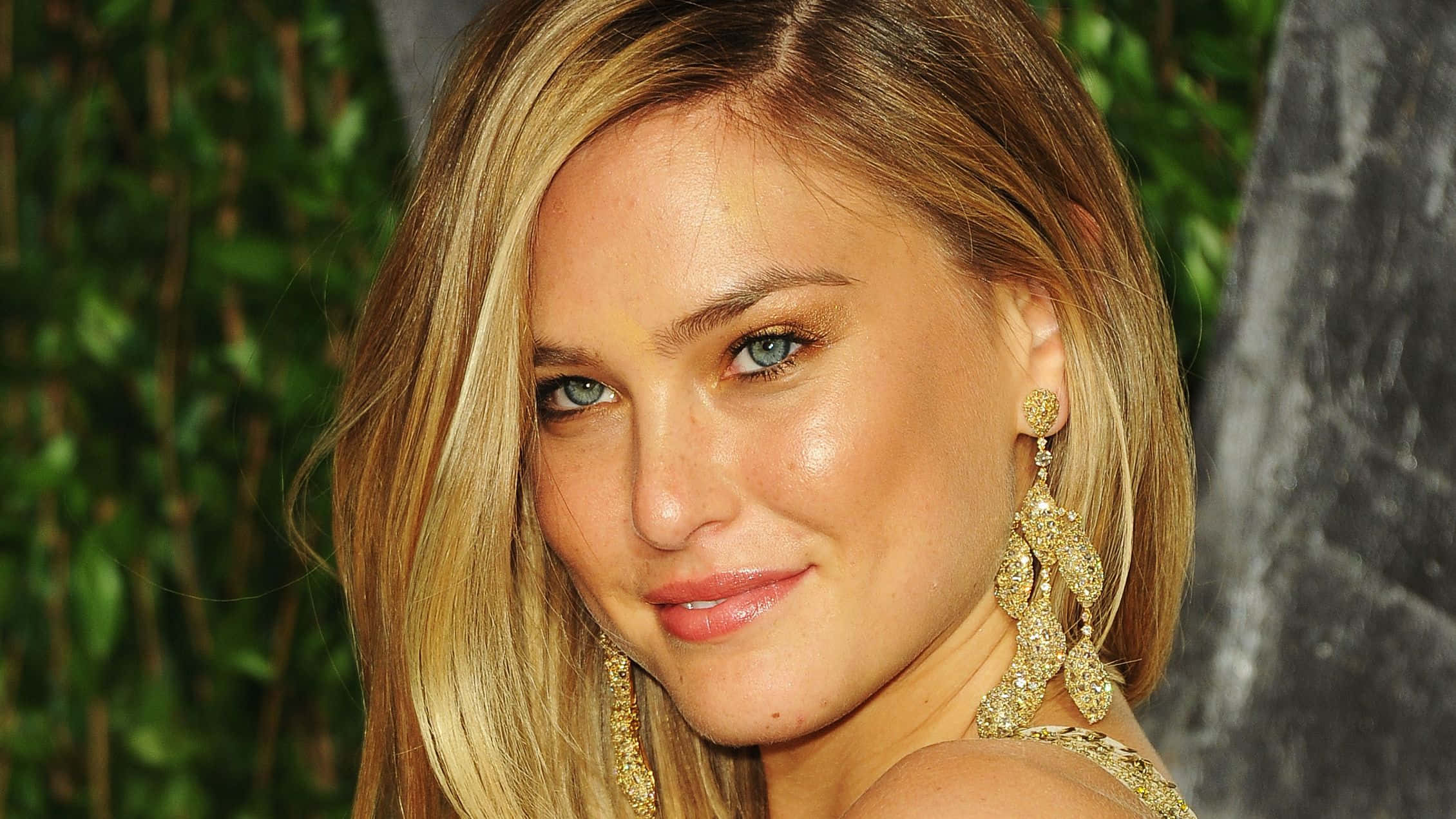 Bar Refaeli 桌布 壁紙 — 一位金髮碧眼、戴著金色垂墜耳環的女性特寫肖像，在模糊的綠色樹葉背景下溫柔微笑。