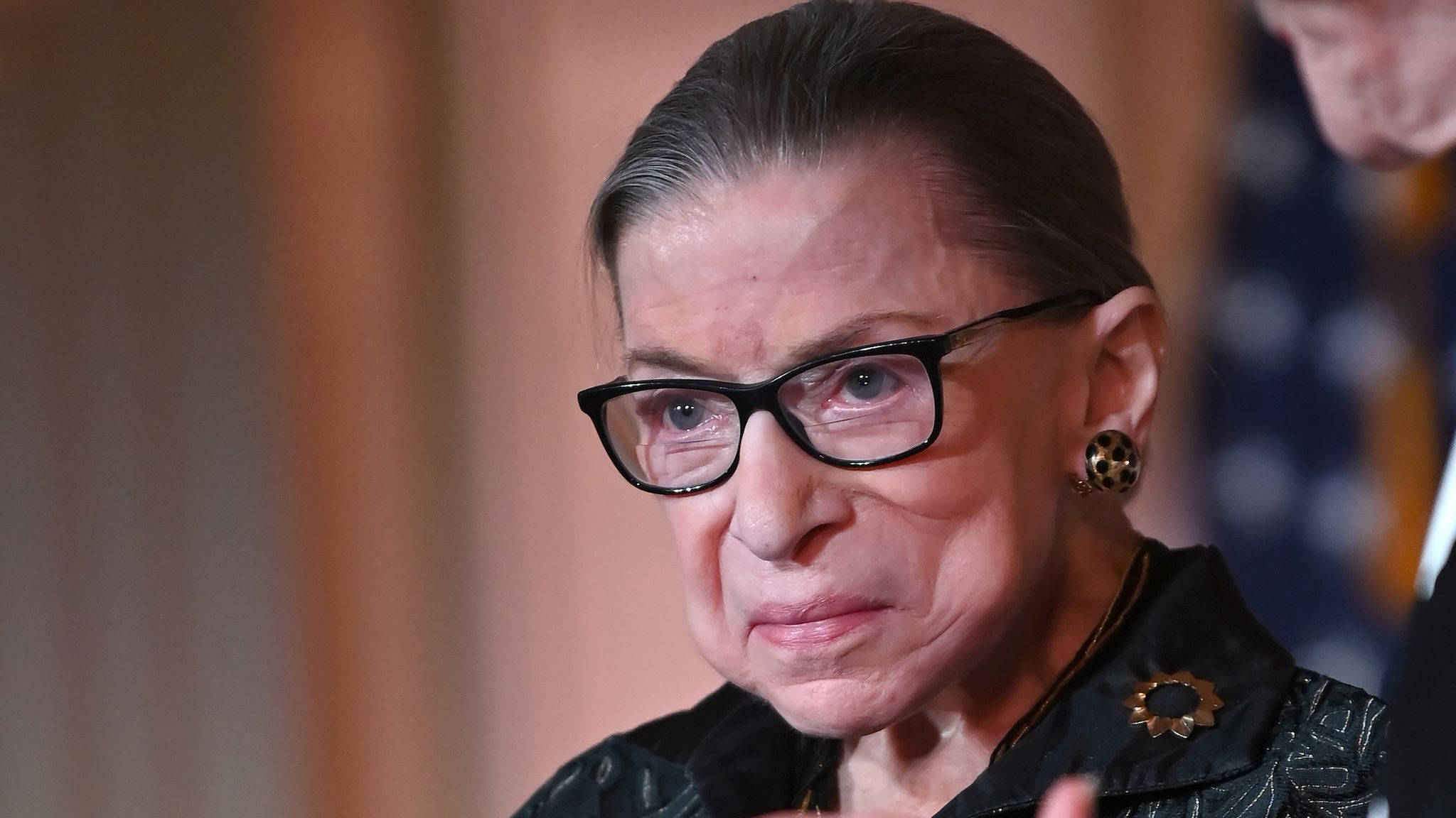 露絲·拜德·金斯伯格 桌布 壁紙 — Ruth Bader Ginsburg 的特寫肖像，戴著黑色框眼鏡和深色圖案外套，上面有個太陽光線胸針。