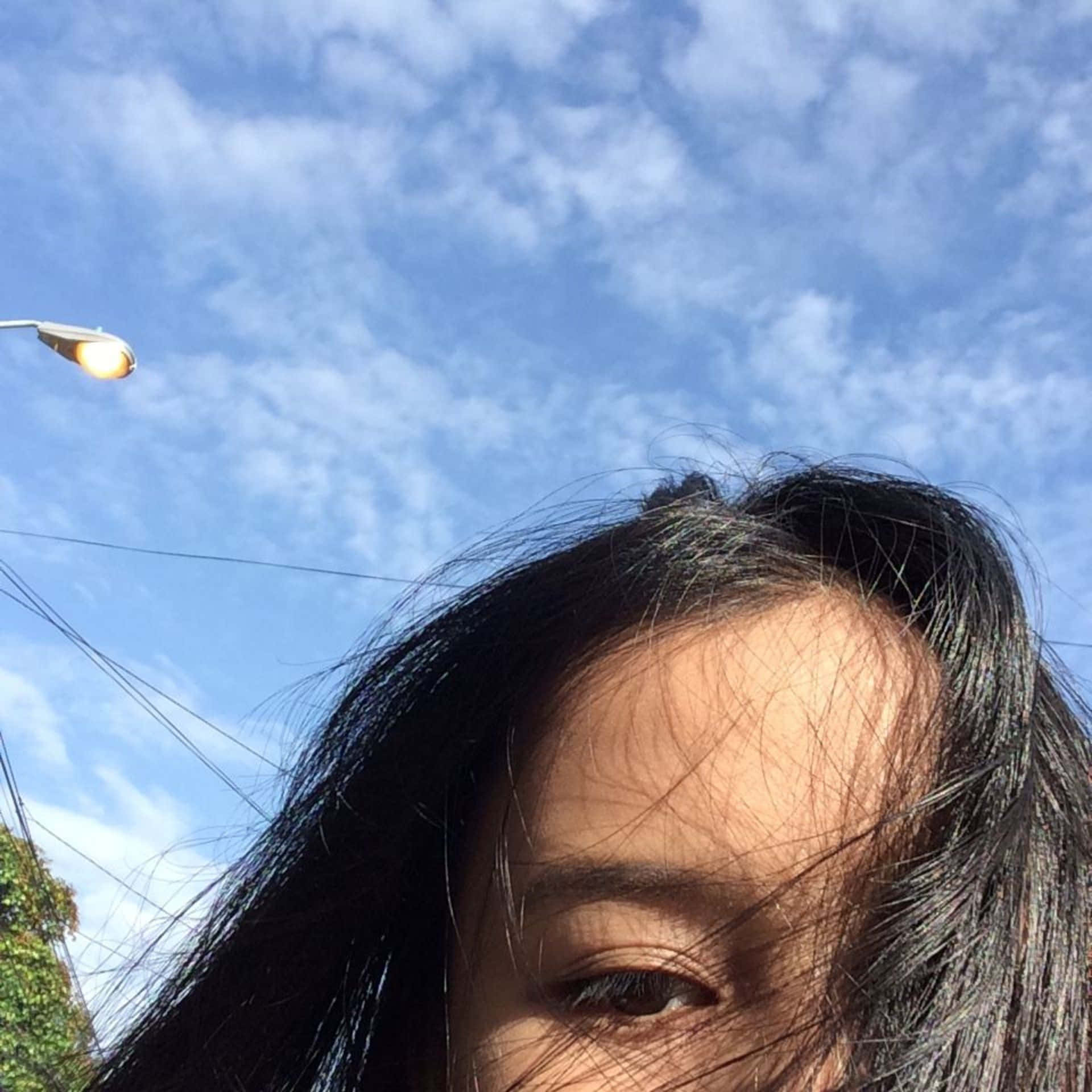  壁紙 — 一個深色頭髮的人的特寫自拍，背景是晴朗藍天，有散雲和街燈。