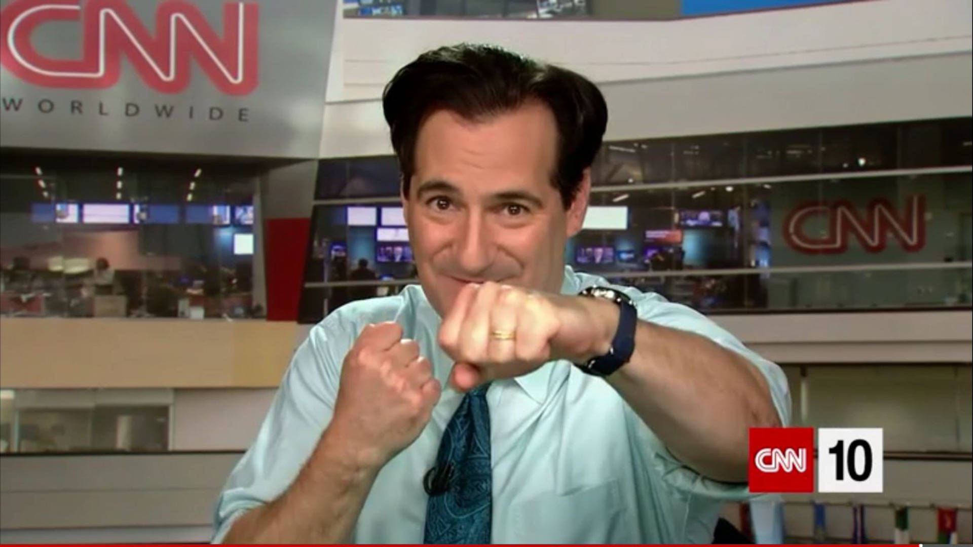 tv 桌布 壁紙 — CNN 攝影棚內一位微笑的新聞主播向鏡頭做出一個玩樂式的拳頭碰撞手勢。
