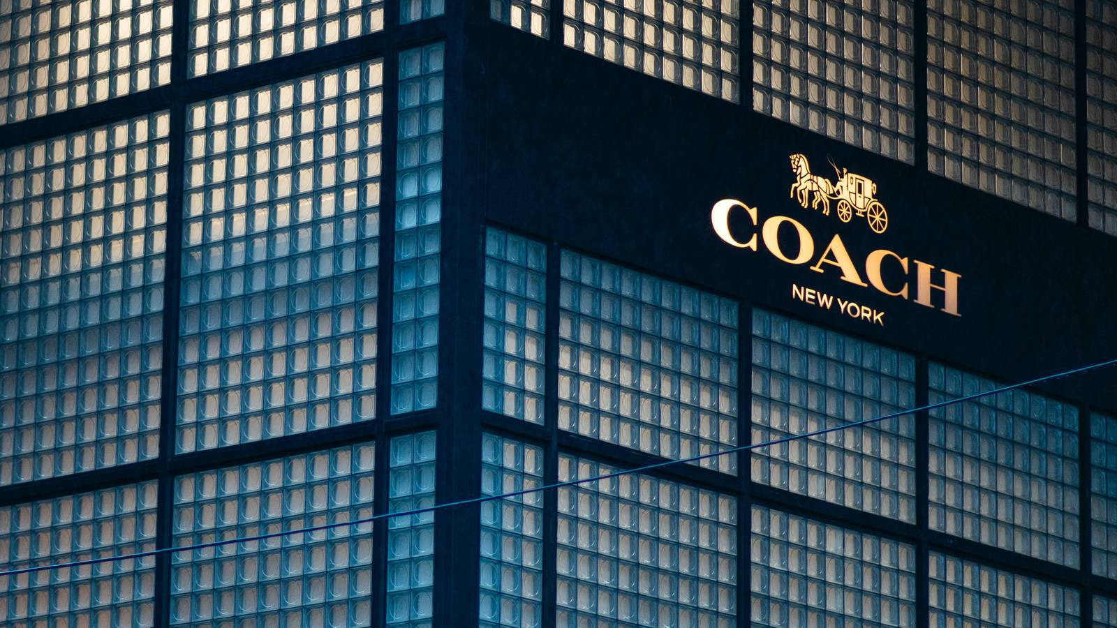 Coach 桌布 壁紙 — Coach New York 商店招牌在現代玻璃鋼建築外牆上閃耀金色的特寫。