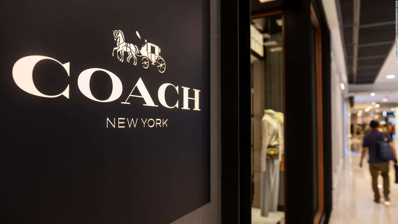 教練 桌布 壁紙 — Coach New York 店鋪標誌的特寫，帶有馬車標誌，背景為深色，後面是模糊的購物中心走廊。