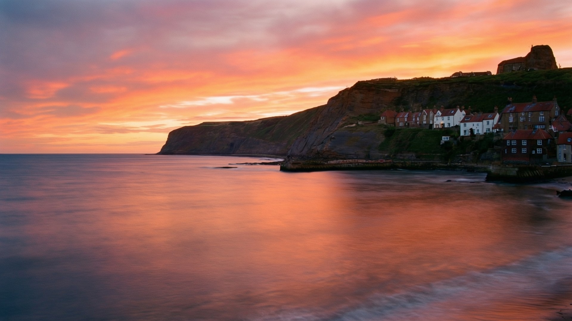 Robin Hood's Bay Sunset 壁紙 — 一個沿海村莊在日落時依附在懸崖邊，熾熱的橙色和粉紅色雲彩倒映在平靜的海面上。