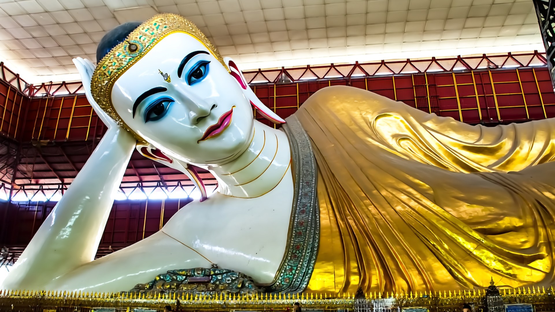 Reclining Buddha Chauk Htat Gyi 壁紙 — 一尊巨大的臥佛，白色皮膚，藍色眼睛，金色長袍，在高高的天花板下安詳地躺著，天花板上有紅色和金色的結構樑。