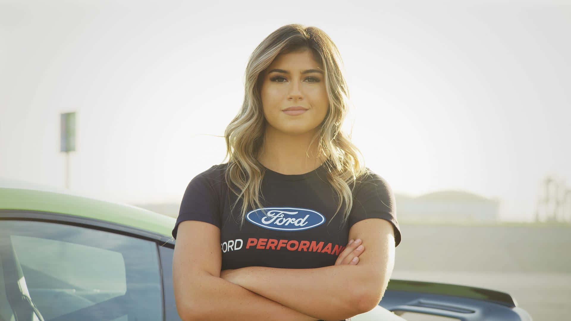  壁紙 — 一位自信的女性穿著Ford Performance襯衫，雙臂交叉，站在一輛時尚的綠色跑車旁，在明亮的日光下。