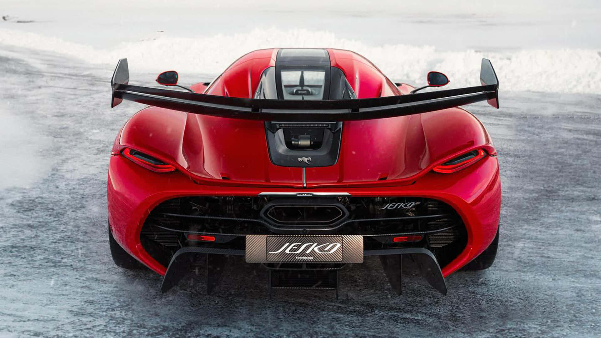  壁紙 — 從後方觀看一輛鮮紅色Koenigsegg Jesko超跑在冰雪覆蓋的道路上行駛，帶有大型黑色尾翼。