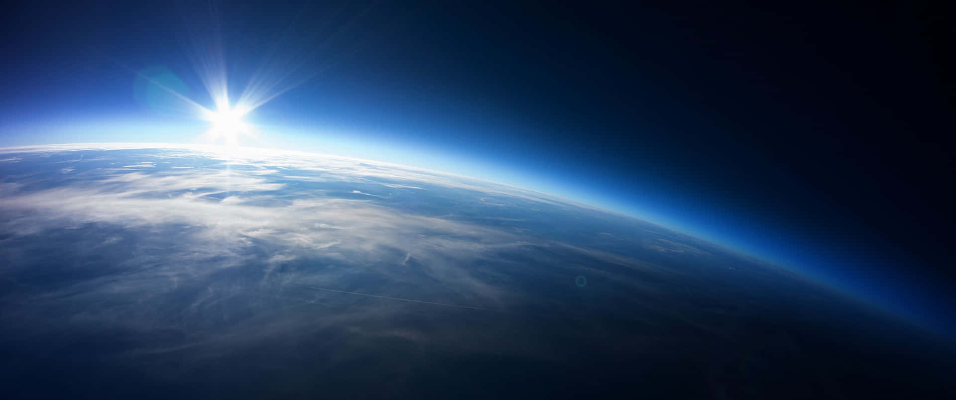 從太空看地球上的日出，顯示藍色大氣層和雲層。 — 3440X1440 壁紙