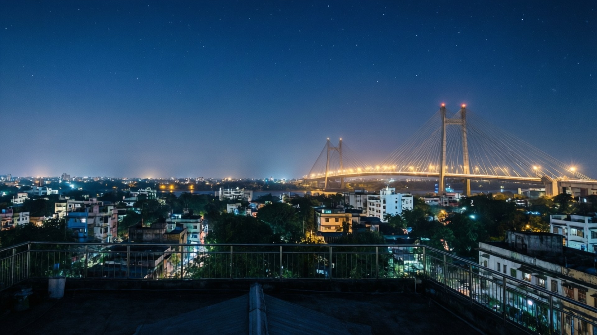 Kolkata Rooftop Vista 壁紙 — 從屋頂俯瞰夜景，顯示一座明亮的吊橋橫跨河流，上方有星空，下方有城市燈光。