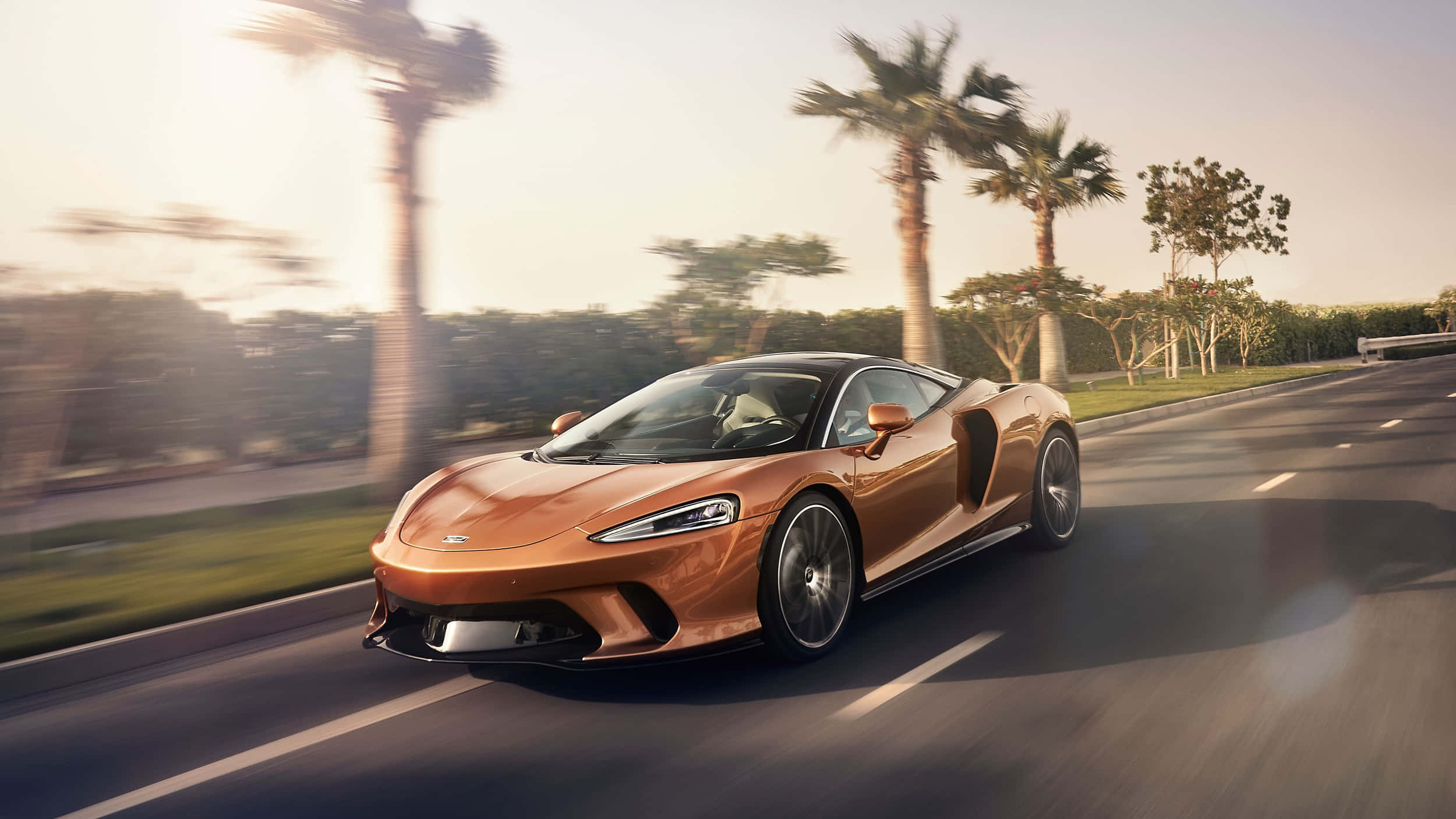 Mclaren Gt 桌布 壁紙 — 一輛銅色McLaren跑車在黃金時段沿著棕櫚樹成蔭的道路疾馳，背景呈現動態模糊效果。