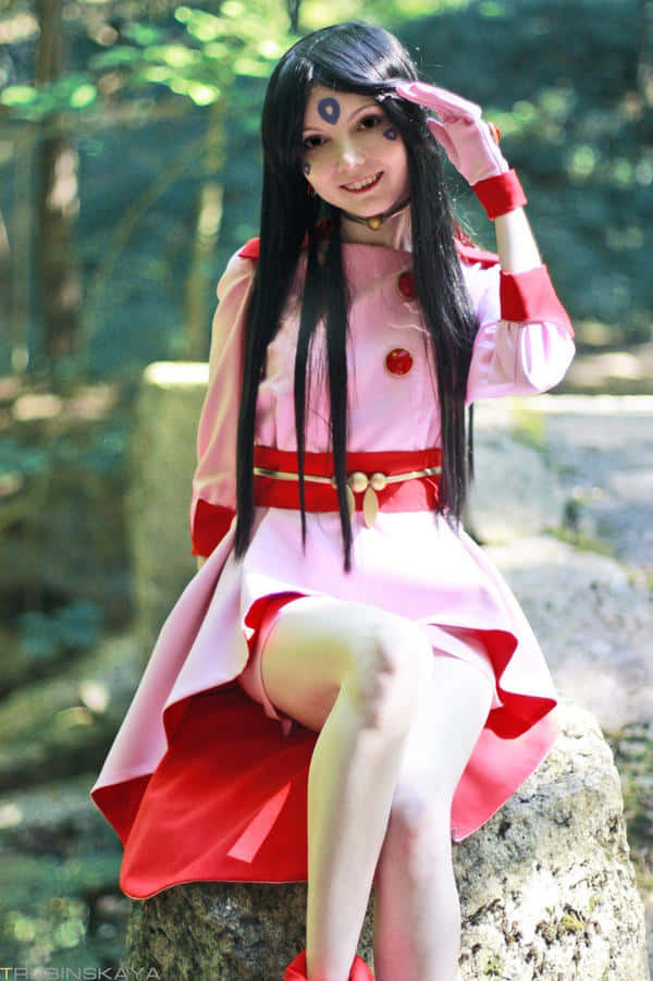  壁紙 — 一位穿著粉紅色和紅色服裝、額頭上有封印的角色扮演者，微笑著坐在陽光普照的森林中長滿苔蘚的石頭上。