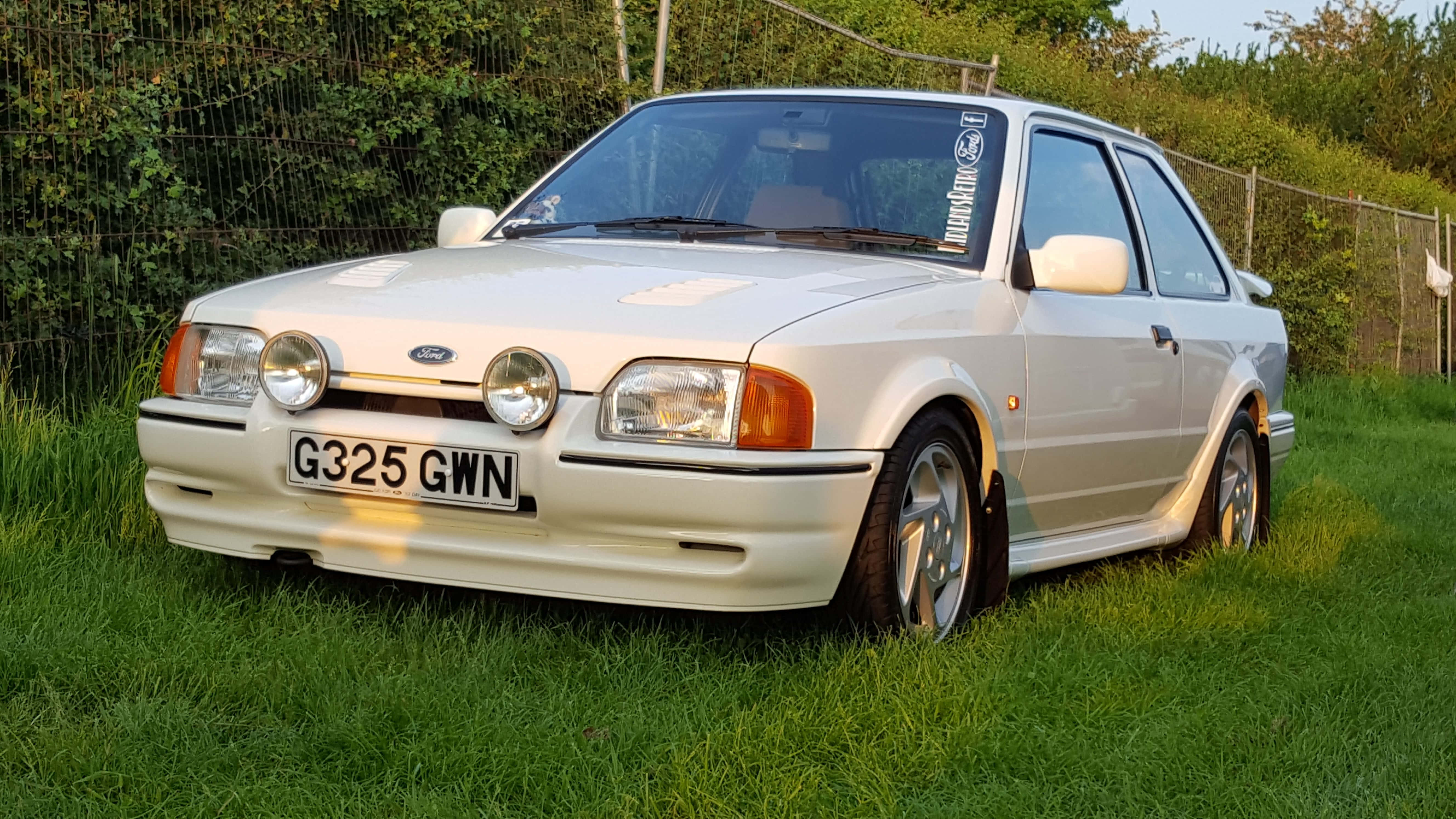 Ford Escort 桌布 壁紙 — 一輛奶油色福特 Sierra Cosworth 停放在草地上，後面有灌木叢和籬笆，在溫暖的傍晚光線下。