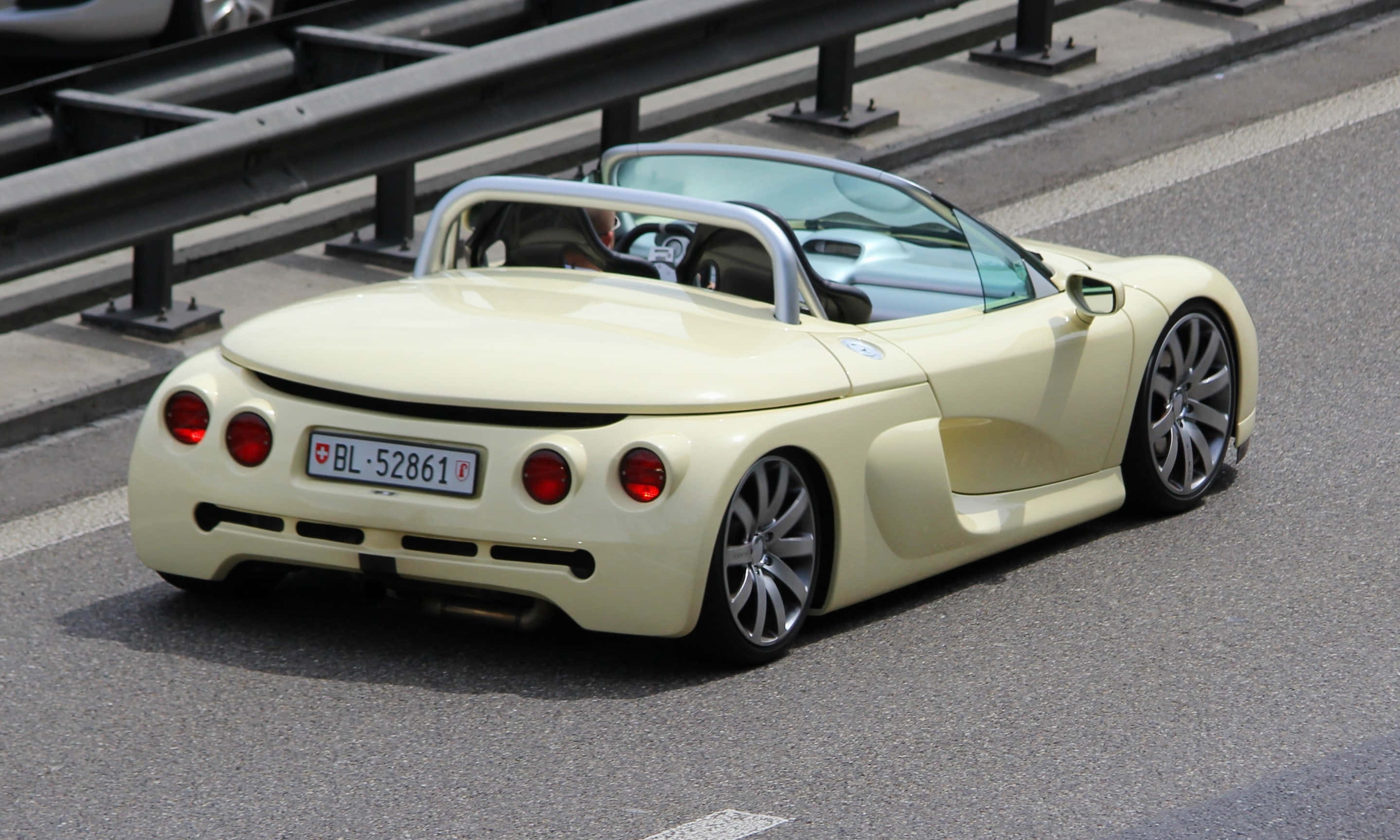 Renault Sport Spider 桌布 壁紙 — 一輛奶油黃色敞篷跑車，帶有紅色尾燈，在陽光普照下，沿著有護欄的高速公路行駛。
