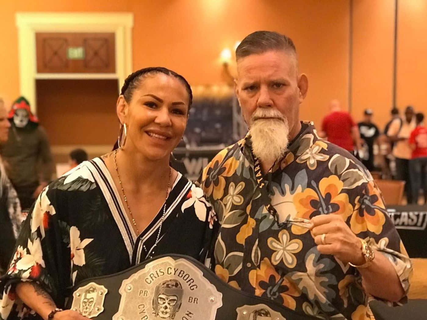  壁紙 — Cris Cyborg 自豪地微笑著，手持冠軍腰帶，與一位穿著花襯衫的鬍鬚男在室內合影。