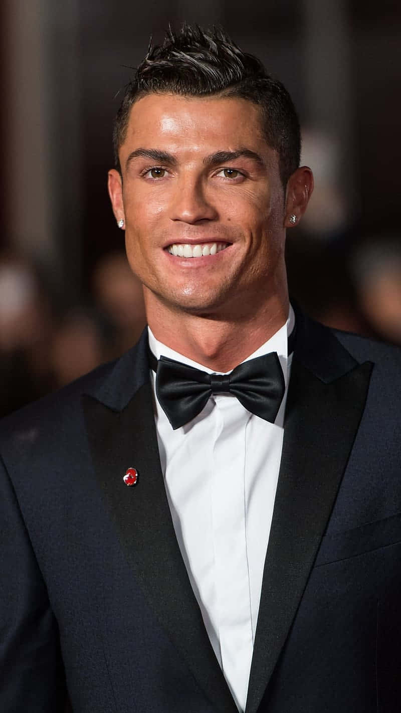  壁紙 — Cristiano Ronaldo 穿著俐落黑色禮服，搭配蝴蝶結和紅色領針，在模糊的活動背景下燦爛微笑。