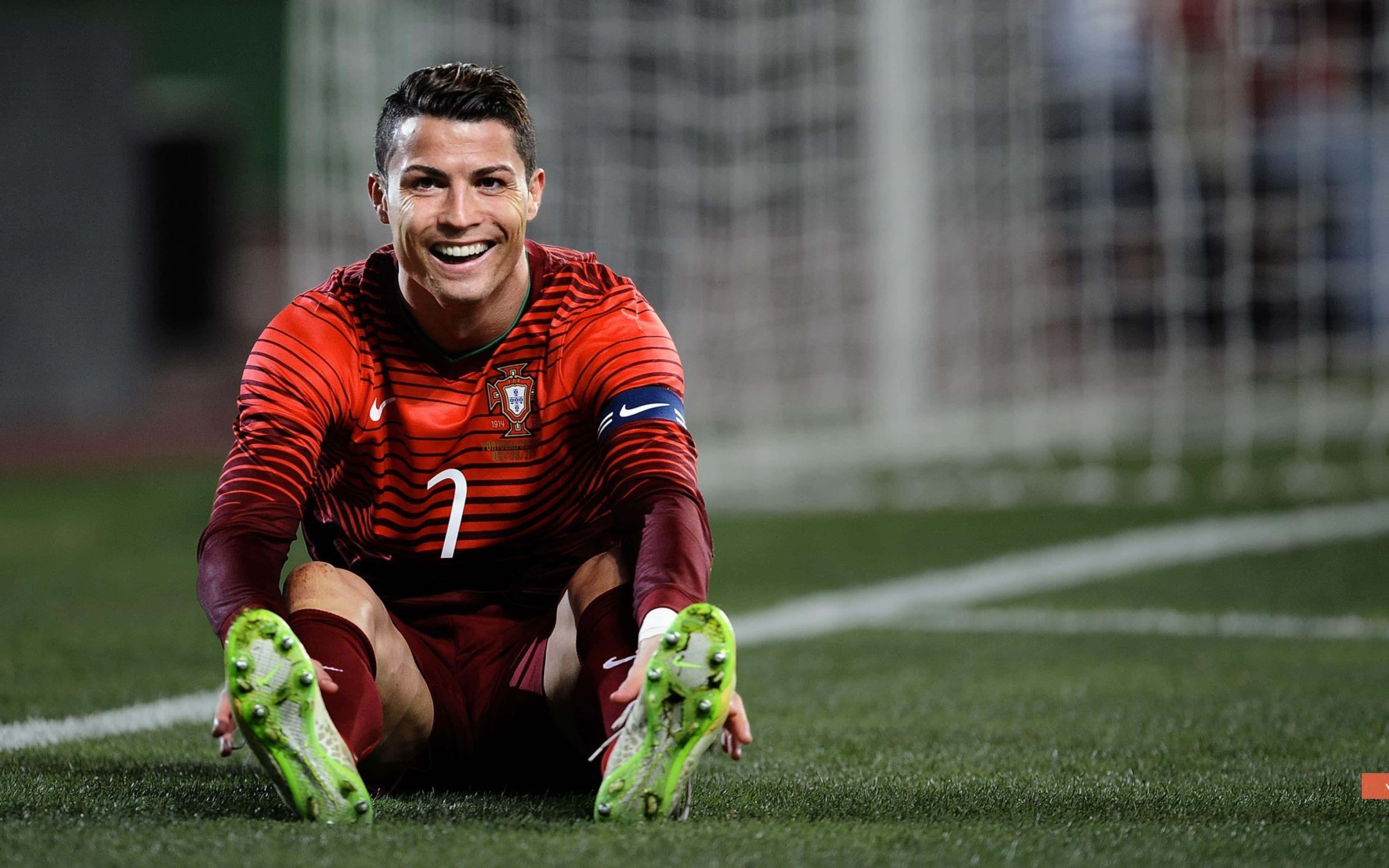Ronaldo 桌布 壁紙 — Cristiano Ronaldo 在綠色足球場上跪下，身穿紅色葡萄牙球衣，露出燦爛的笑容，腳踏亮綠色球鞋。