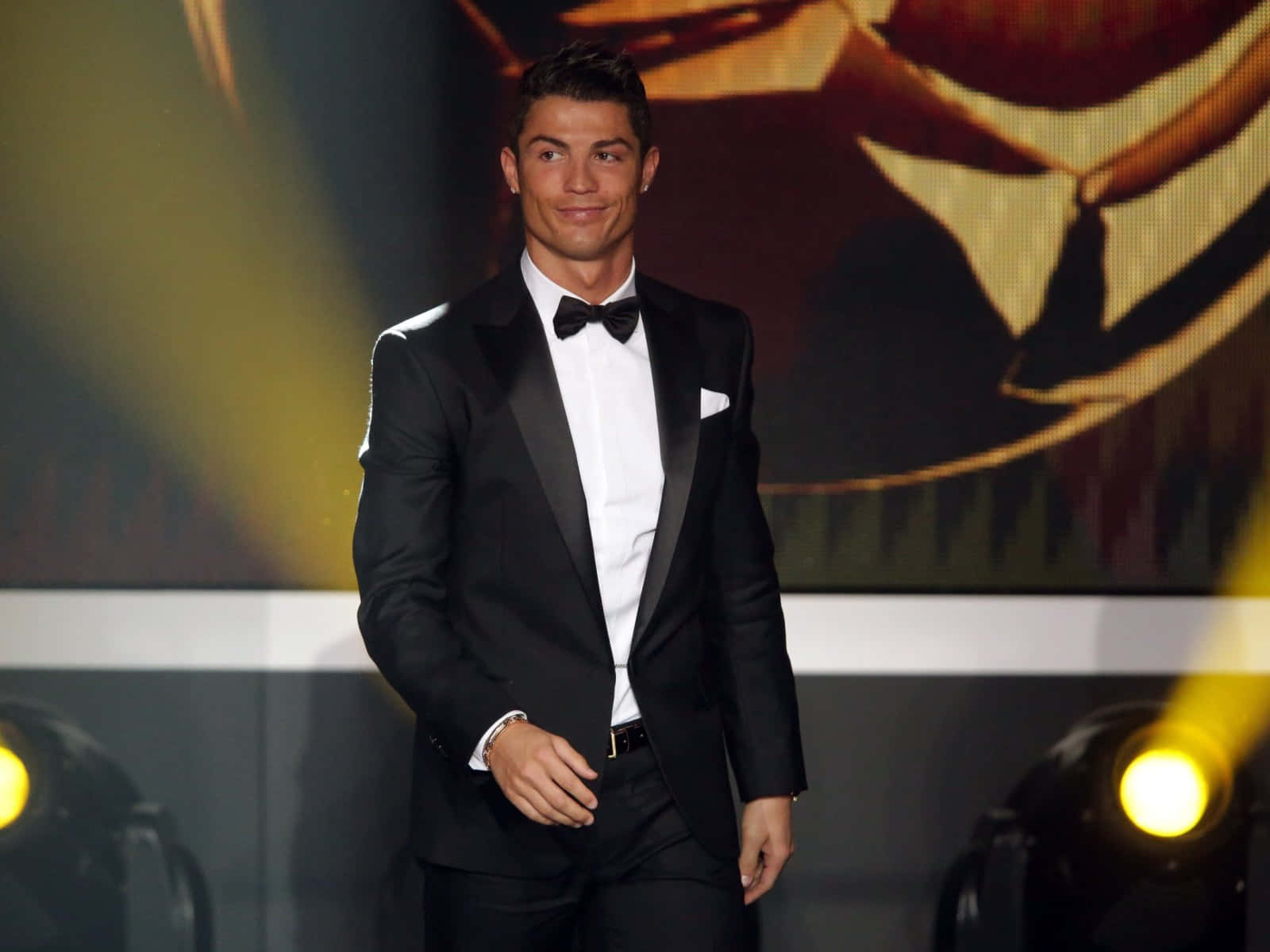  壁紙 — Cristiano Ronaldo 身穿黑色修身燕尾服，在金色聚光燈照耀的明亮舞台上自信微笑。