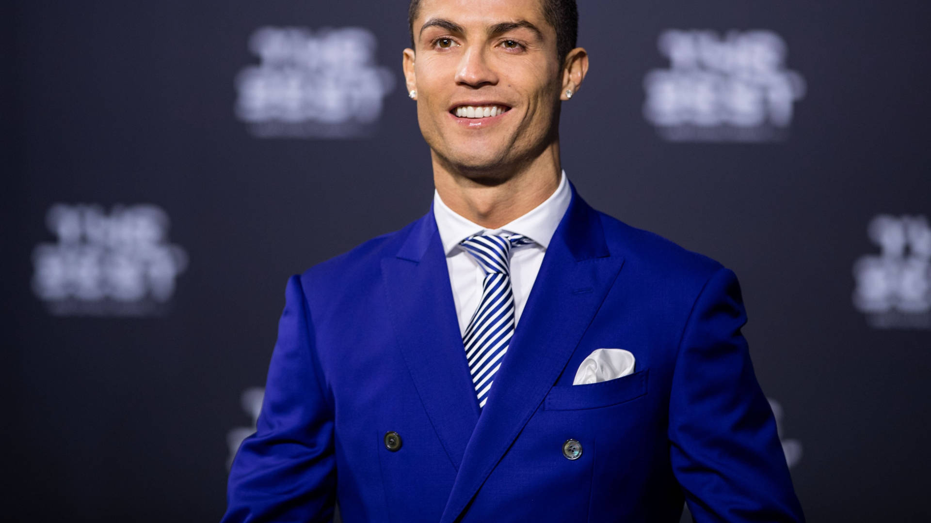 克里斯蒂亞諾·羅納度 桌布 壁紙 — Cristiano Ronaldo 身穿剪裁俐落的皇家藍雙排扣西裝，搭配條紋領帶，在深色品牌背景前自信微笑。