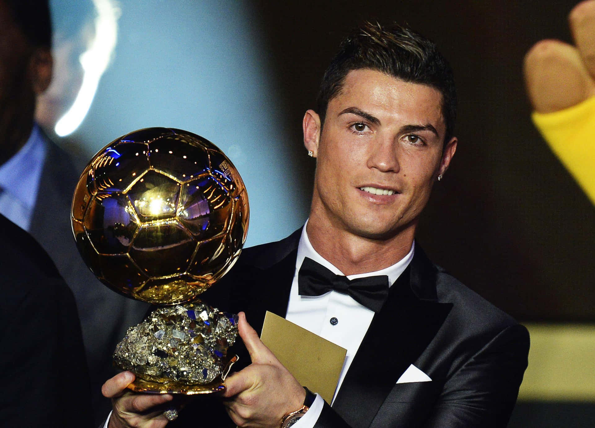  壁紙 — Cristiano Ronaldo 自豪地微笑，手持閃耀的金色足球獎盃，身穿正式黑色燕尾服。