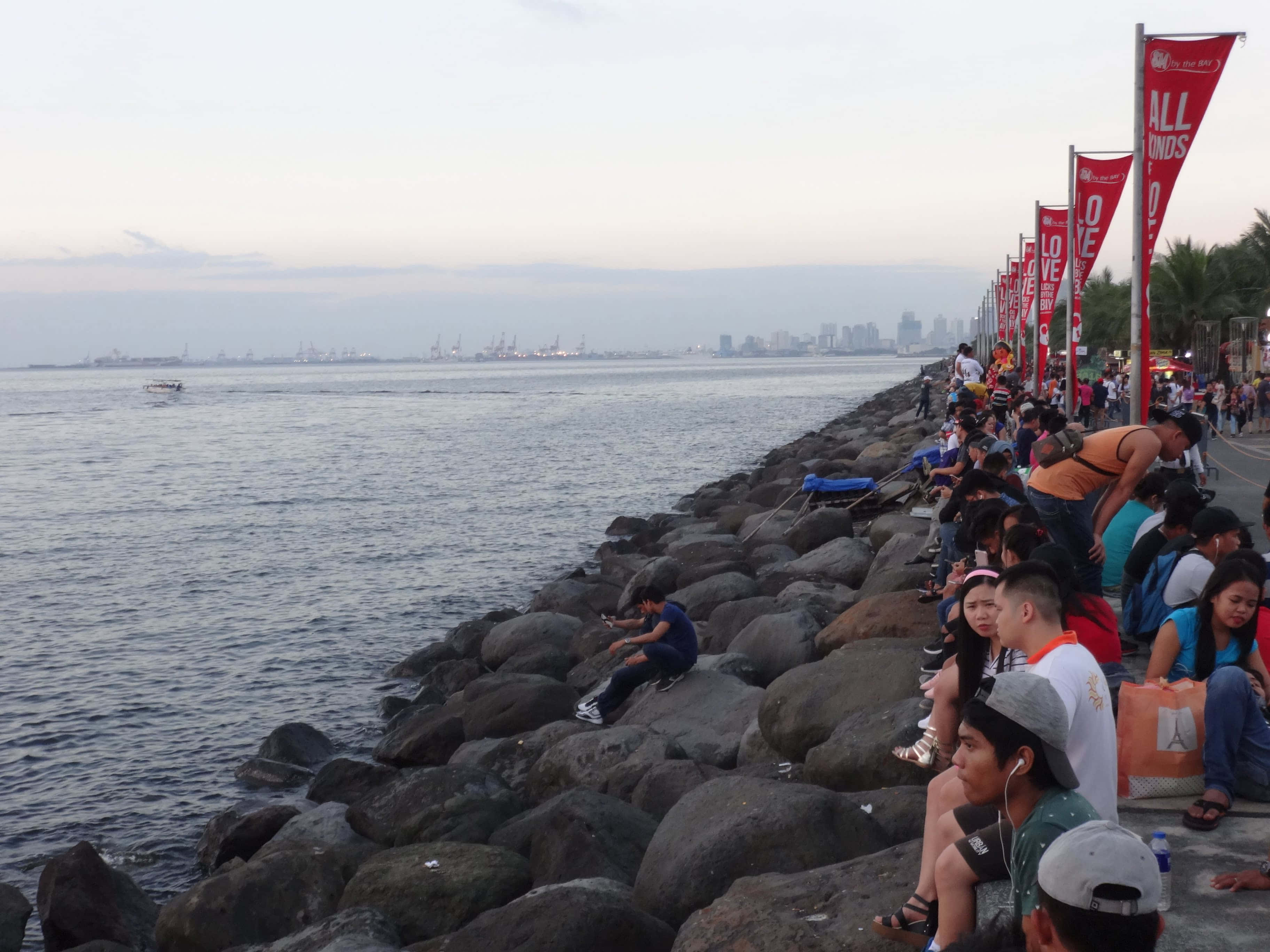 Seaside 桌布 壁紙 — 一群人在黃昏時分沿著岩石海濱放鬆和釣魚，遠處是城市天際線。